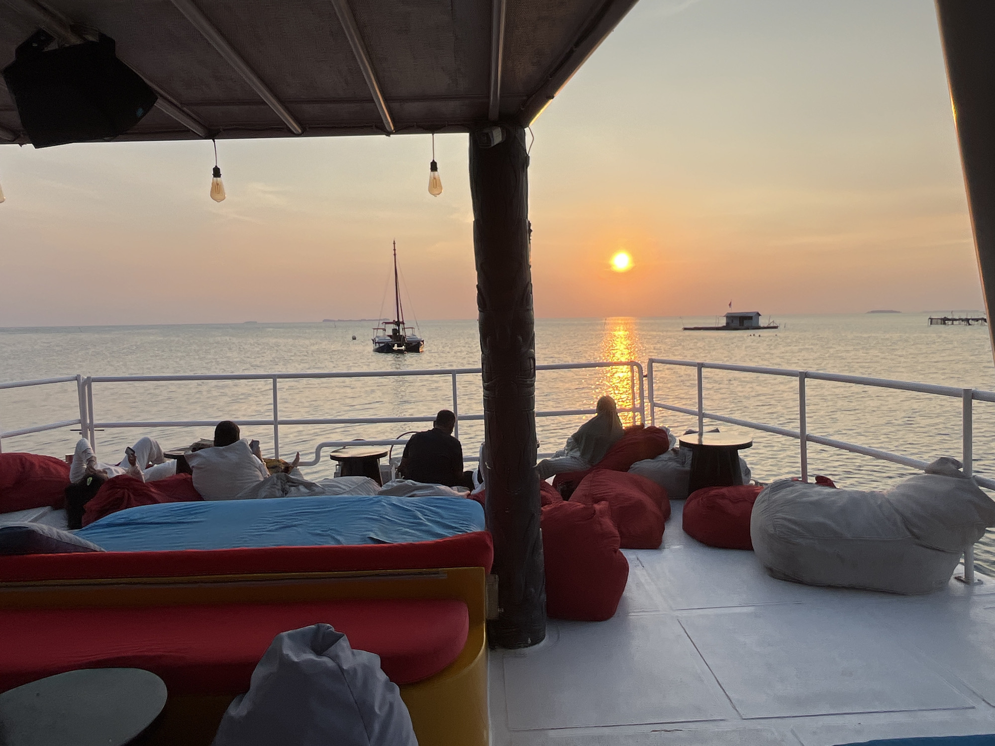 An exterior, eye-level, wide shot shows a covered deck of a boat with people relaxing during sunset. The sun is setting over the ocean, casting a warm glow on the water. The deck is furnished with beanbag chairs and cushions. There are several people on the deck, some sitting and some reclining. In the distance, there are a few boats and a small structure on the water. The sky is a gradient of orange and yellow, with a few clouds visible. The water is calm and reflects the colors of the sunset. The scene appears to be peaceful and serene. The image was taken in Karimunjawa, Indonesia.