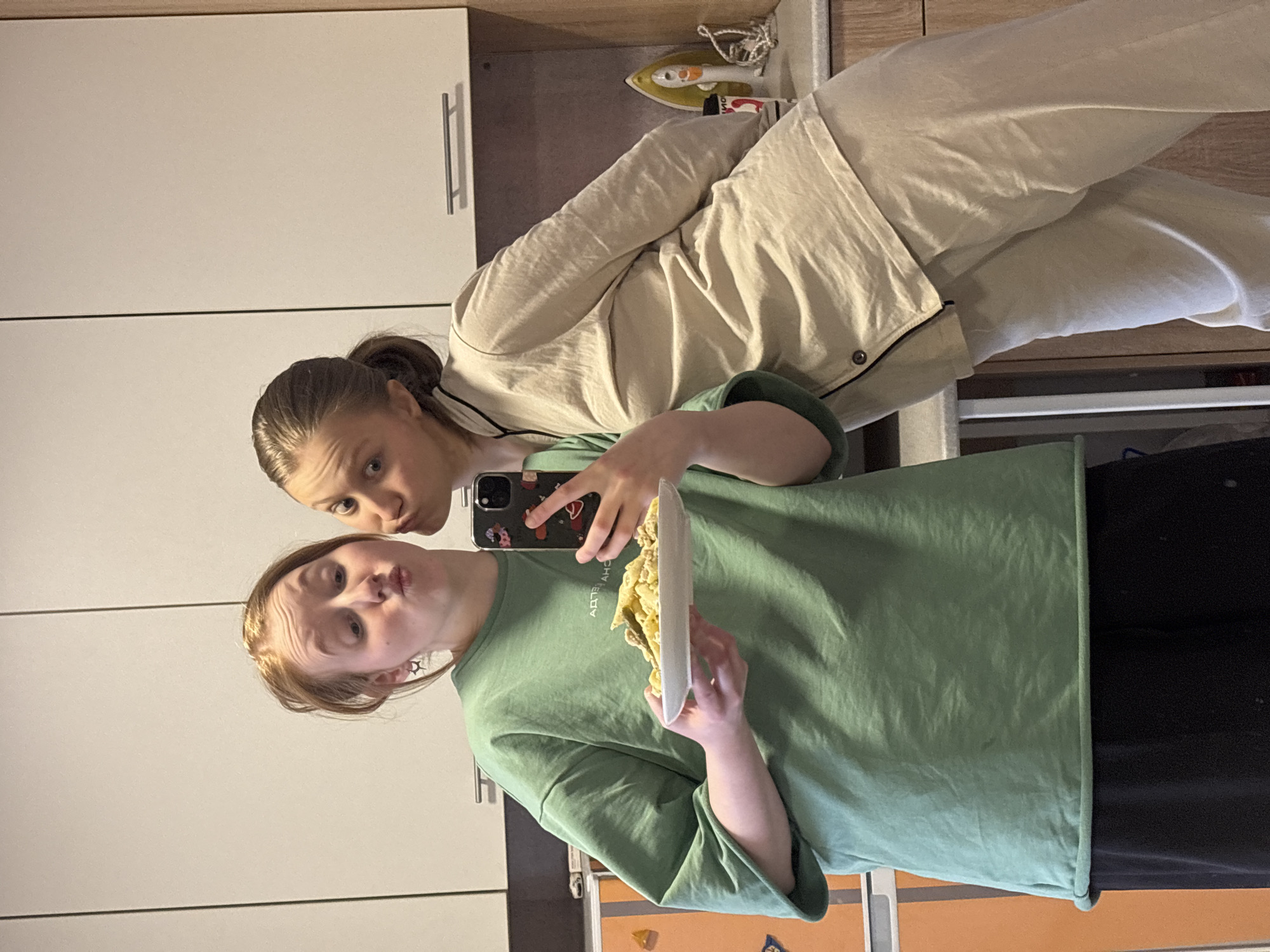 Two females are taking a selfie in a kitchen setting. One is holding a plate of food and the other is holding a smartphone. The kitchen features white cabinets and drawers, a wooden countertop, and an iron on top of the counter. The location is likely within the city of Perm, Russia.