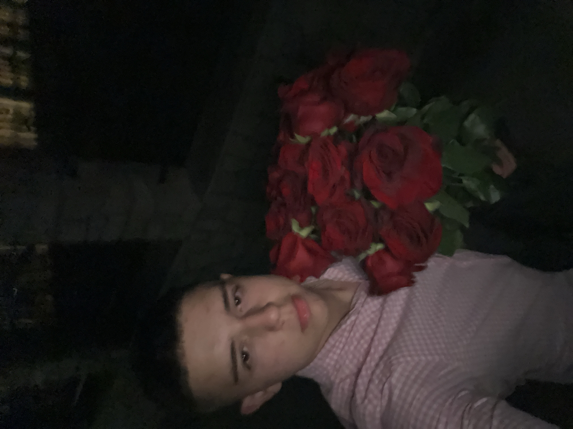A young male individual is lying down, with a bouquet of red roses placed on his chest. The image is taken in low lighting conditions. The background is dark and obscured, but it appears to be outdoors. Based on the location of Zlatograd, Bulgaria, the location may be a public space in the city. The man is wearing a pink and white checkered shirt. His facial expression is neutral.