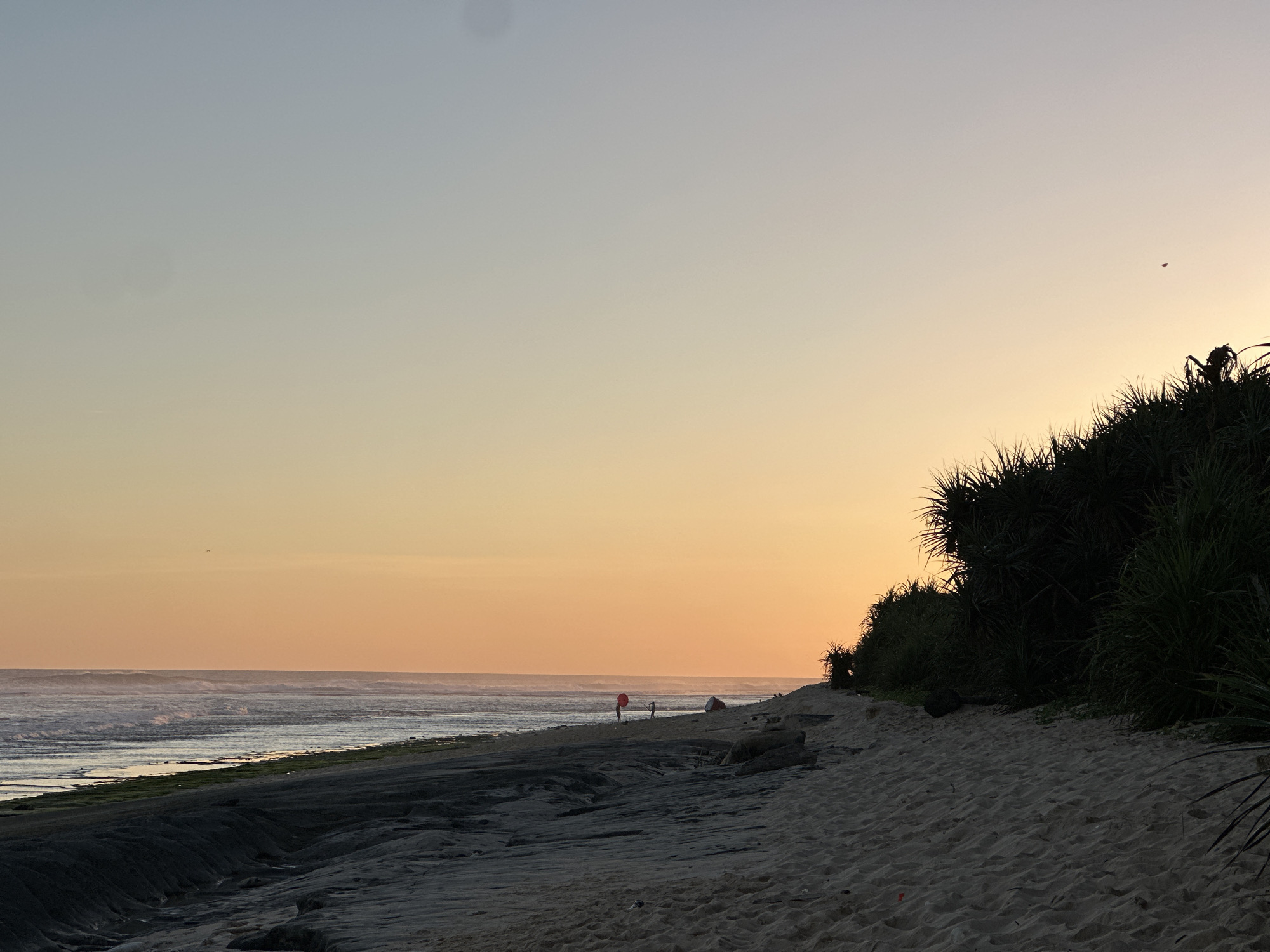The photograph depicts a beach in Kuta Selatan, Indonesia, at dusk. The sky transitions from light blue to orange near the horizon. The ocean has waves breaking along the shore. Three people are visible in the distance on the beach. Two of them are near a red object, possibly a balloon or flag. The shoreline has dark sand near the water and light sand further inland, with vegetation covering a small dune on the right side of the frame.