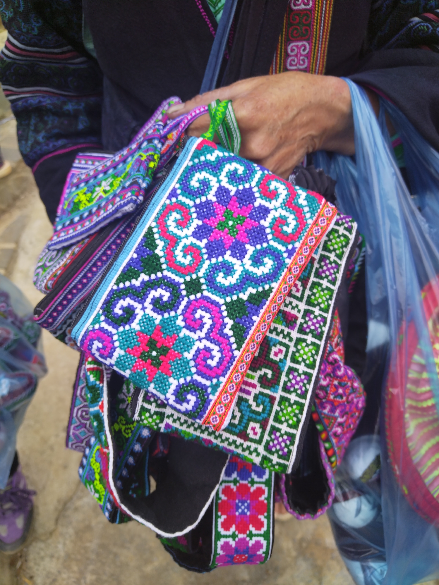 A person stands outdoors in an undefined urban setting, holding a collection of intricately patterned, colorful bags. The bags are adorned with geometric and floral designs in hues of blue, green, pink, white, and purple. The person's hand, visibly aged with prominent veins, grasps a green woven handle of one of the bags. The person is wearing a dark-colored shirt or dress with detailed embroidery around the collar and sleeves. A partially visible blue plastic bag is also held by the person. The ground below is gray, and a blurred pair of purple shoes is visible in the lower-left corner of the image. The overall lighting is soft and diffused.