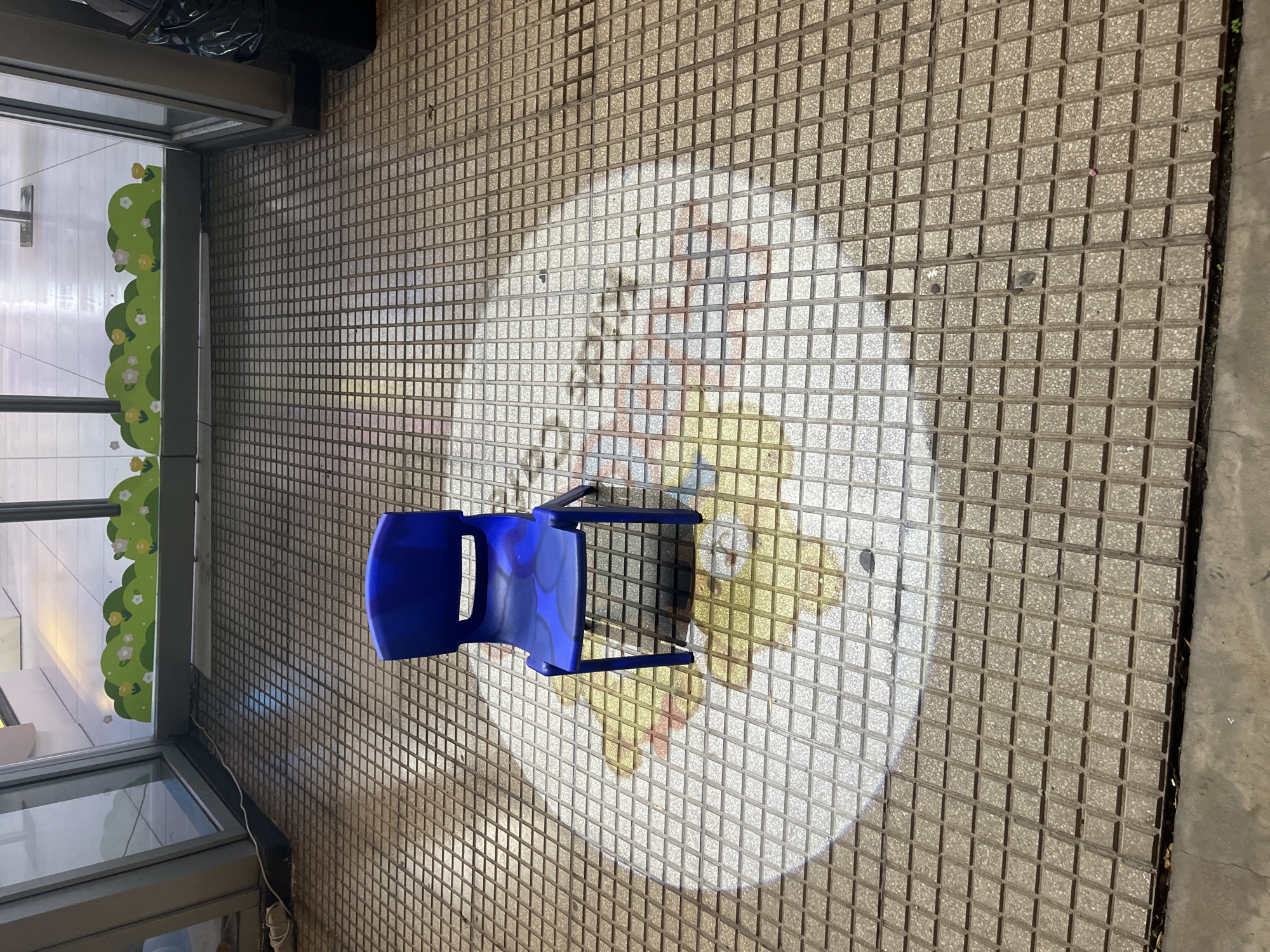 The image shows a blue plastic chair on a tiled floor. The floor is composed of gray square tiles with darker grout. A circular design is visible on the floor under the chair, seemingly painted or applied to the tiles. The chair appears to be in an indoor setting near a window with decorative floral accents. A trash bag is partially visible in the upper left corner of the image. The location is presumed to be in Buenos Aires, Argentina, but no evidence is found to confirm or deny the city.