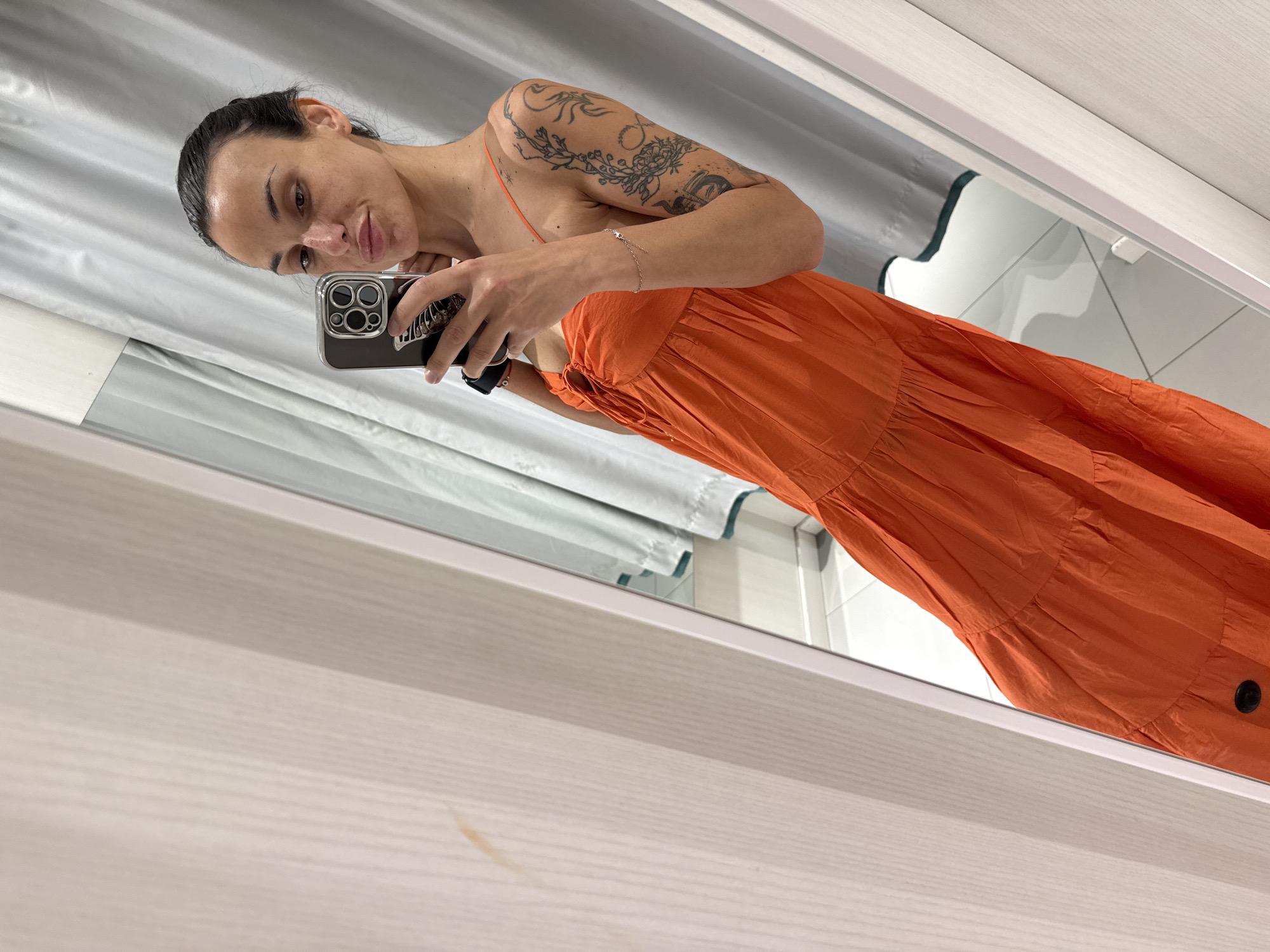 A woman with dark hair pulled back in a bun, wearing an orange dress, stands in a fitting room and takes a selfie with her phone. She has tattoos on her left arm. The fitting room has light-colored walls and gray curtains.