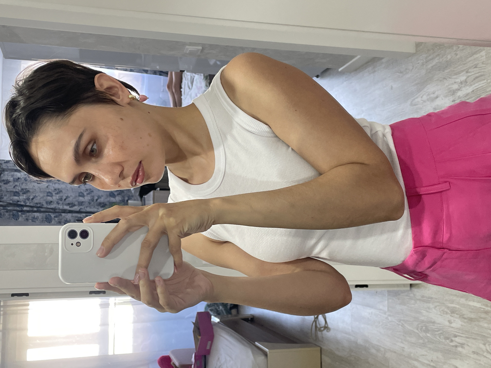 A woman is taking a selfie in a room with a mirror. She is wearing a white tank top and pink pants. She is holding a white smartphone in her hands. The background shows a window with blinds, a bed, and a section of the room's interior. The location in the city of Izhevsk, Russia, is not identifiable from the image.