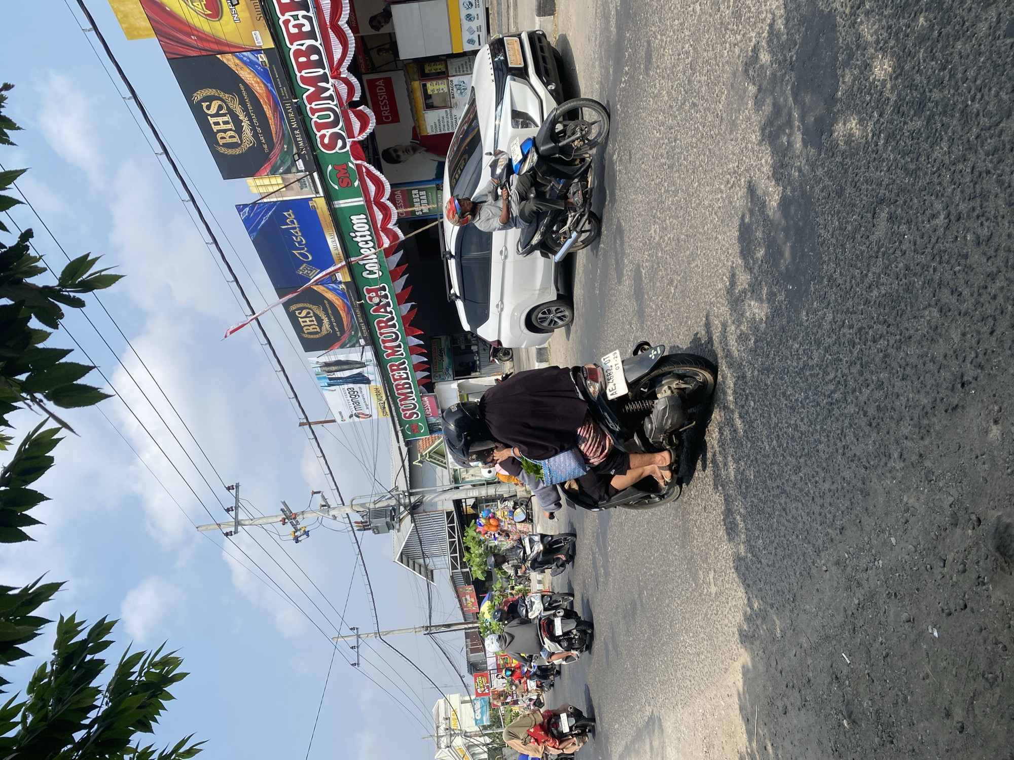 The image depicts a street scene in Sempol, Indonesia, likely a commercial area, on a bright, sunny day. A busy street is filled with motorcycles, with a few cars also present. Several individuals are riding motorcycles, some with passengers. One motorcycle in the foreground has a rider wearing a helmet and carrying a basket. Across the street, a white SUV is parked. Buildings line the street, featuring advertisements for various businesses, including "BHS" and "Sumber Murah Collection." Overhead, power lines and utility poles are visible, along with green foliage from trees on the left side of the frame. The road surface appears to be asphalt. The overall atmosphere suggests a typical day in a bustling Indonesian town.