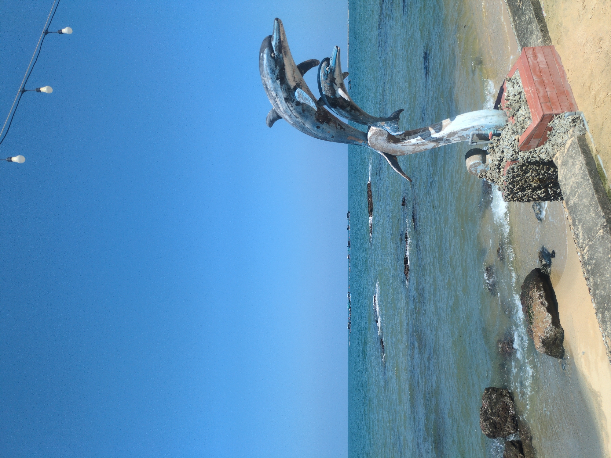 The image shows a statue of dolphins on the shore of Phú Quốc, Vietnam. The statue depicts three dolphins leaping out of the water. The statue is near the water and a concrete wall, with a sandy beach next to it. The ocean is visible in the background, with scattered rocks and boats farther out. The sky is clear blue. Overhead are light bulbs strung on a cable.