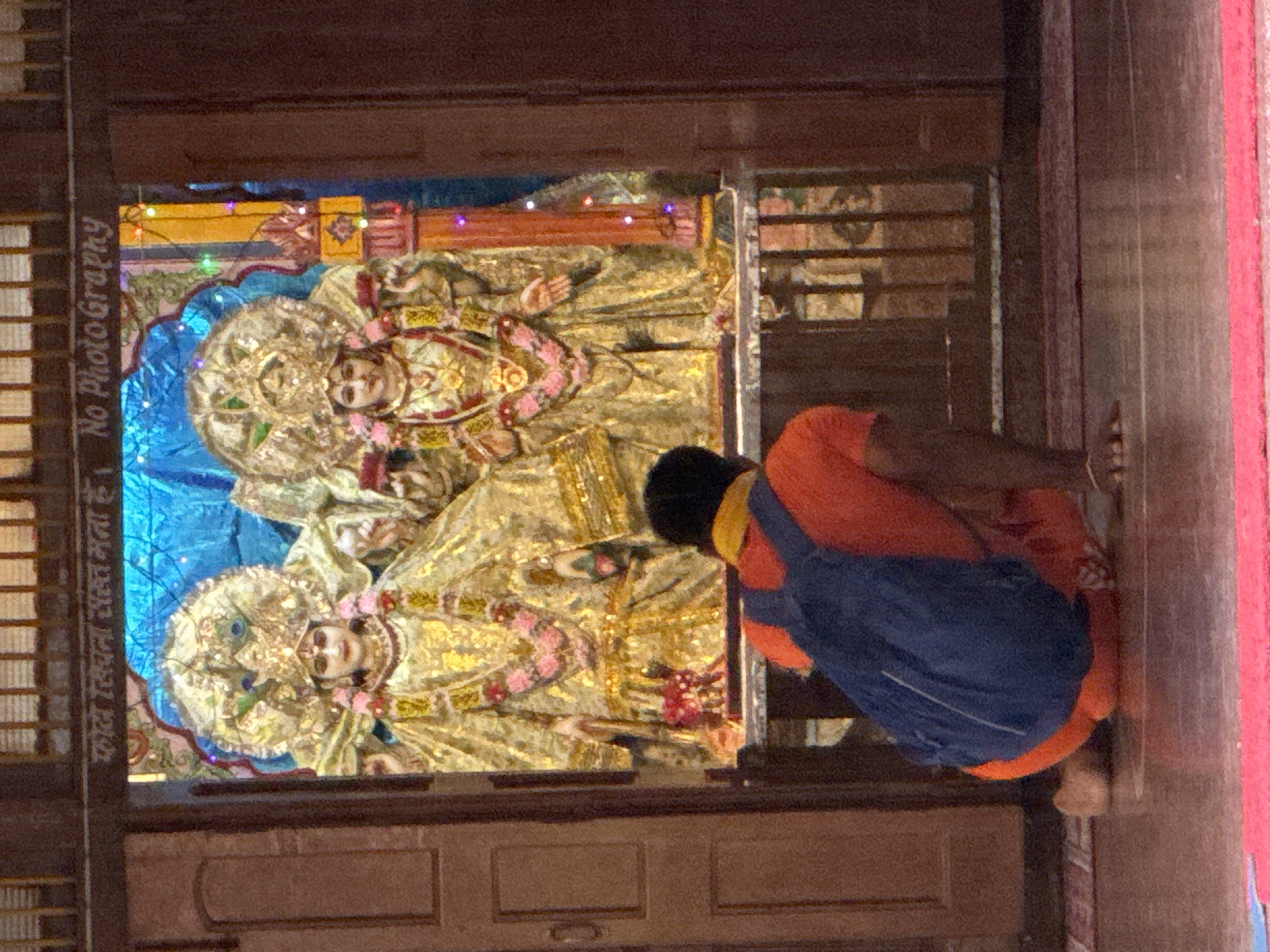In Rishikesh, India, at the entrance of a temple, a person wearing an orange shirt and a blue backpack kneels on the floor, facing two elaborately dressed statues of deities. The statues, adorned with golden attire, crowns, and floral garlands, are positioned within an alcove decorated with blue fabric and colorful lights. Above the alcove, text in Hindi is visible, along with the English words "No Photography." The overall environment suggests a place of worship, with wooden doors flanking the entrance and a tiled floor.