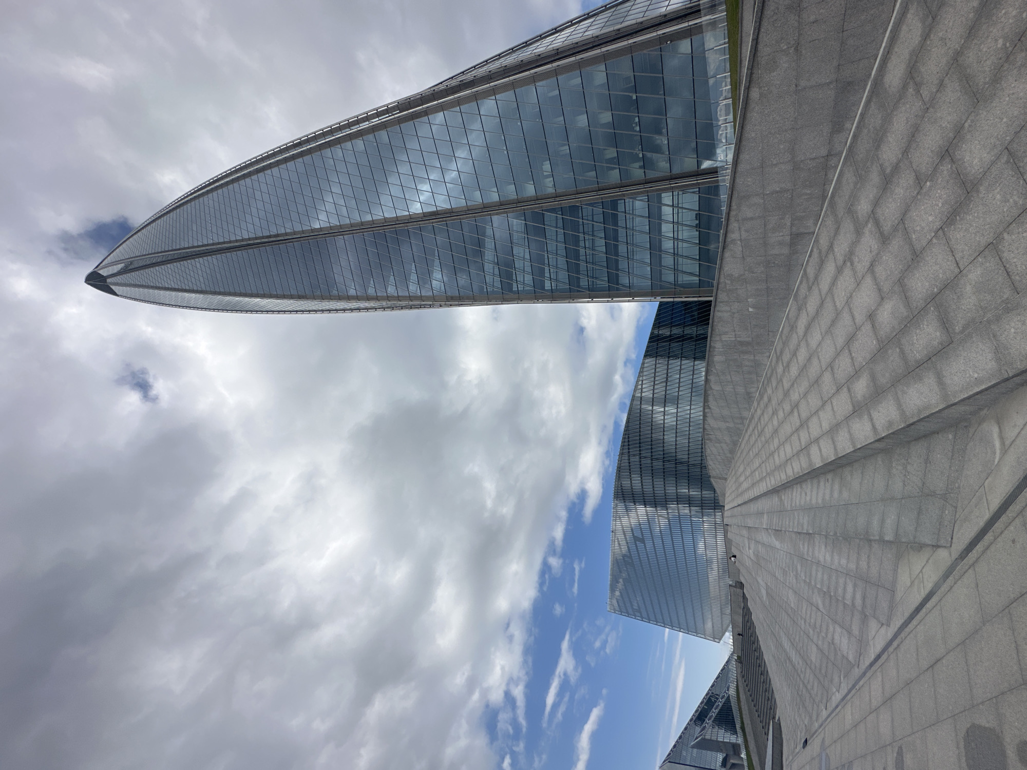 The image depicts the Lakhta Center in Saint Petersburg, Russia, a prominent skyscraper with a distinctive curved, tapering design. The building's facade is composed of glass panels that reflect the cloudy sky, creating a dramatic visual effect. In the foreground, a wide expanse of stone paving forms a sloping plaza with tiered steps leading up towards the buildings. The plaza is clean and well-maintained. Two individuals are visible in the distance, one sitting on the steps and another walking further up the plaza. The sky is overcast with patches of blue showing through, suggesting a dynamic weather condition. Other modern buildings with glass facades are partially visible in the background, contributing to the contemporary architectural landscape.