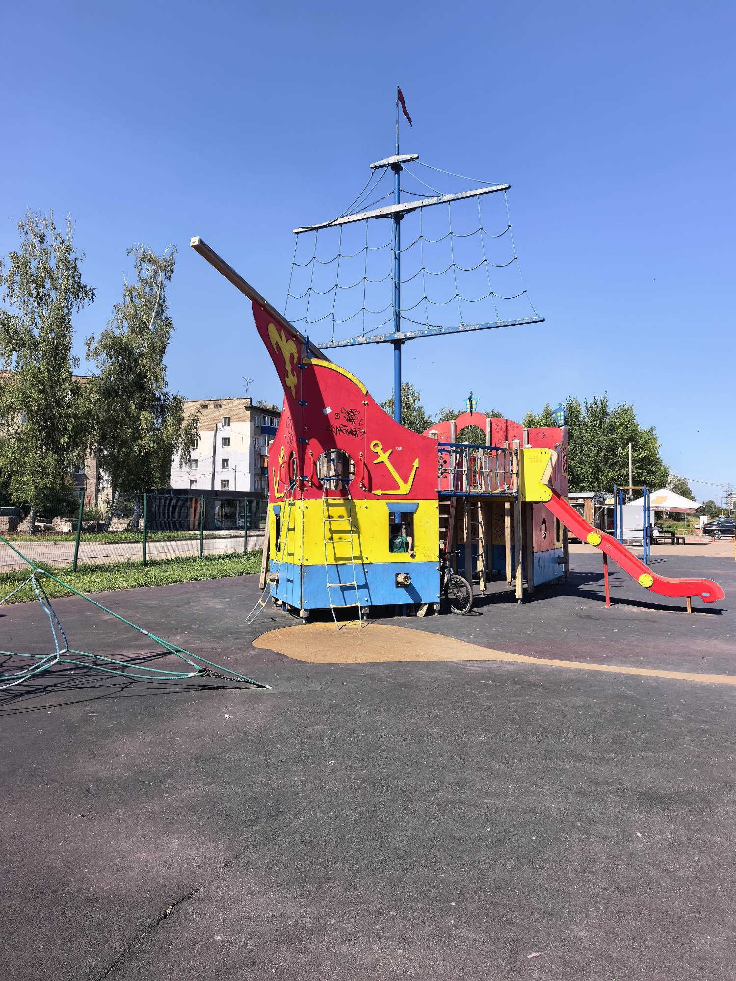A brightly colored pirate ship-themed playground structure stands on a black asphalt surface under a clear blue sky. The playground is predominantly red, yellow, and blue, with a prominent red slide extending from the main structure. A bicycle is parked near the base of the structure. In the background, trees and multi-story buildings are visible, and a fence lines the edge of the playground area. The overall scene suggests a public park or recreational area within the city of Sterlitamak, Russia.