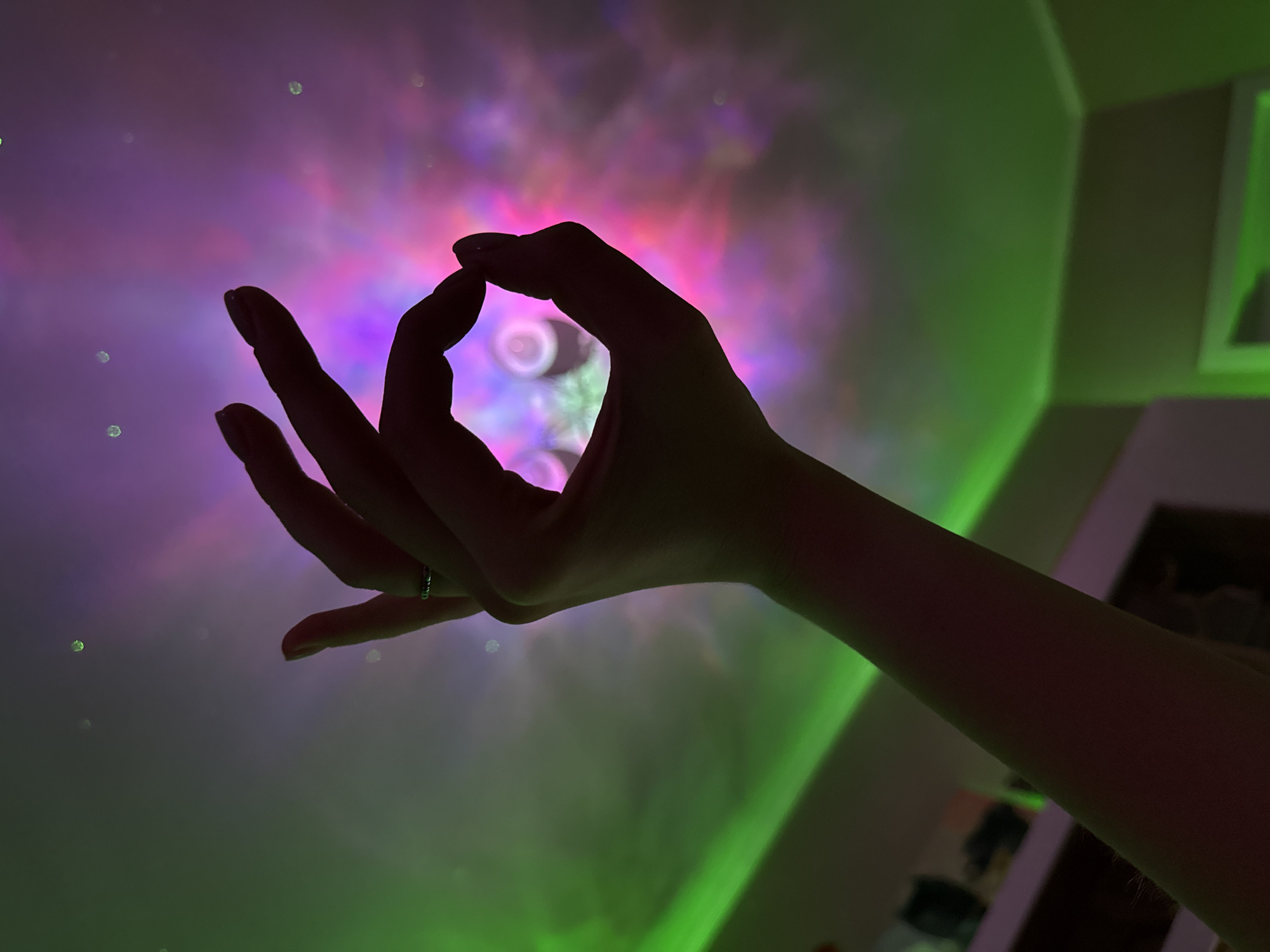 A silhouetted hand forms an "OK" sign against a ceiling illuminated by a nebula projector. The projector casts a gradient of green and pink light, with small white sparkles, creating a celestial effect. The hand, with a ring on the ring finger, is positioned in the foreground. The background shows the corner of a room with shelves and possibly a doorway, also lit by the projector's effects.