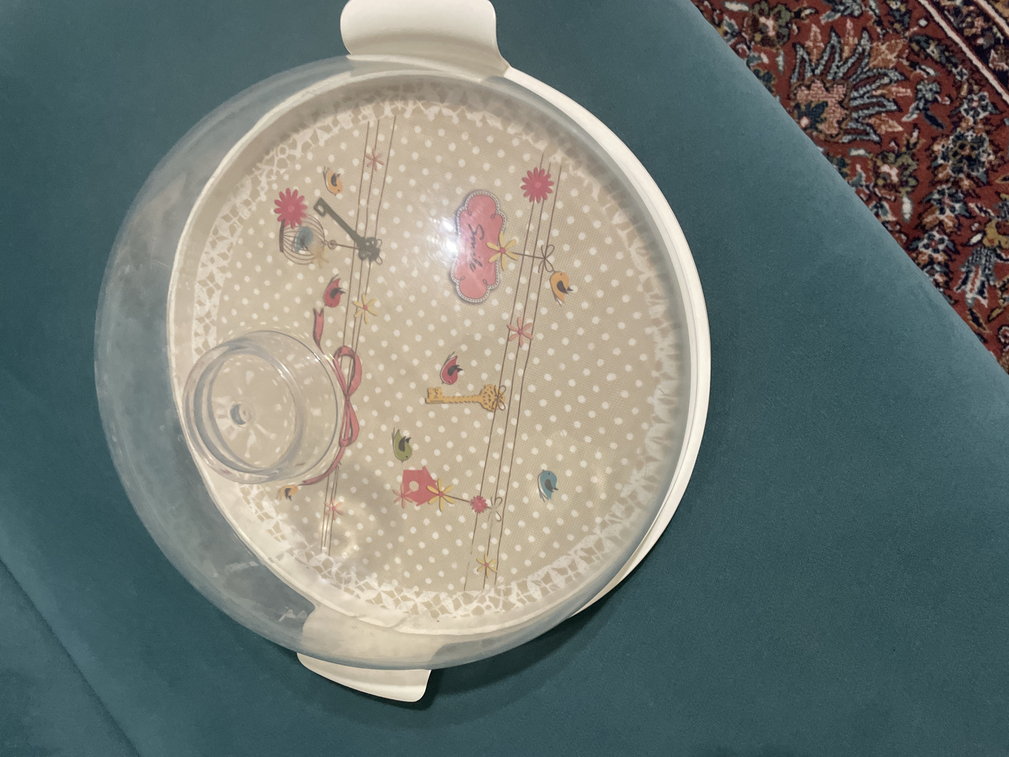 A clear plastic dome lid with white handles sits atop a round cake stand. The cake stand has a light beige base with a subtle pattern of white polka dots and a decorative lace-like edge. Printed on the cake stand are various whimsical illustrations including a birdhouse on a string, small birds, flowers, a key, and a cloud-shaped speech bubble with the word "smile" written in cursive and a pink bow. The dome lid is slightly raised, revealing the interior of the cake stand. The cake stand is placed on a dark teal-colored surface, which appears to be a sofa or upholstered furniture. In the lower right corner of the image, a portion of a patterned rug with red, blue, and green motifs is visible. The scene is indoors, and there are no people or animals present. The lighting is soft and diffused, suggesting an indoor setting with ambient light. The location in Kadıköy, Turkey is not discernible from the image.