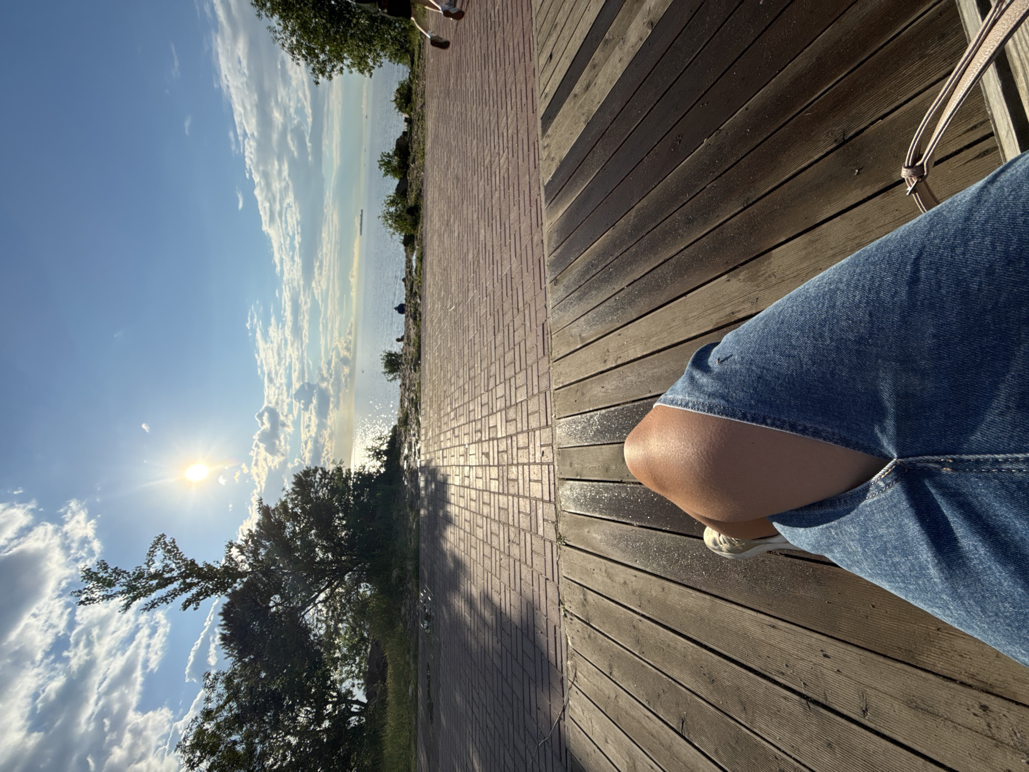 The image captures a person's legs, clad in denim jeans and a sneaker, resting on a wooden deck. The deck is illuminated by sunlight, casting shadows. Beyond the deck is a brick walkway with dappled shade from trees. In the background, a body of water stretches to the horizon, reflecting the bright sun and dotted with clouds. The scene appears to be an outdoor public space, possibly a park or promenade, situated in a city. The time of day is indicated by the low angle of the sun and the warm light, suggesting late afternoon or early morning.