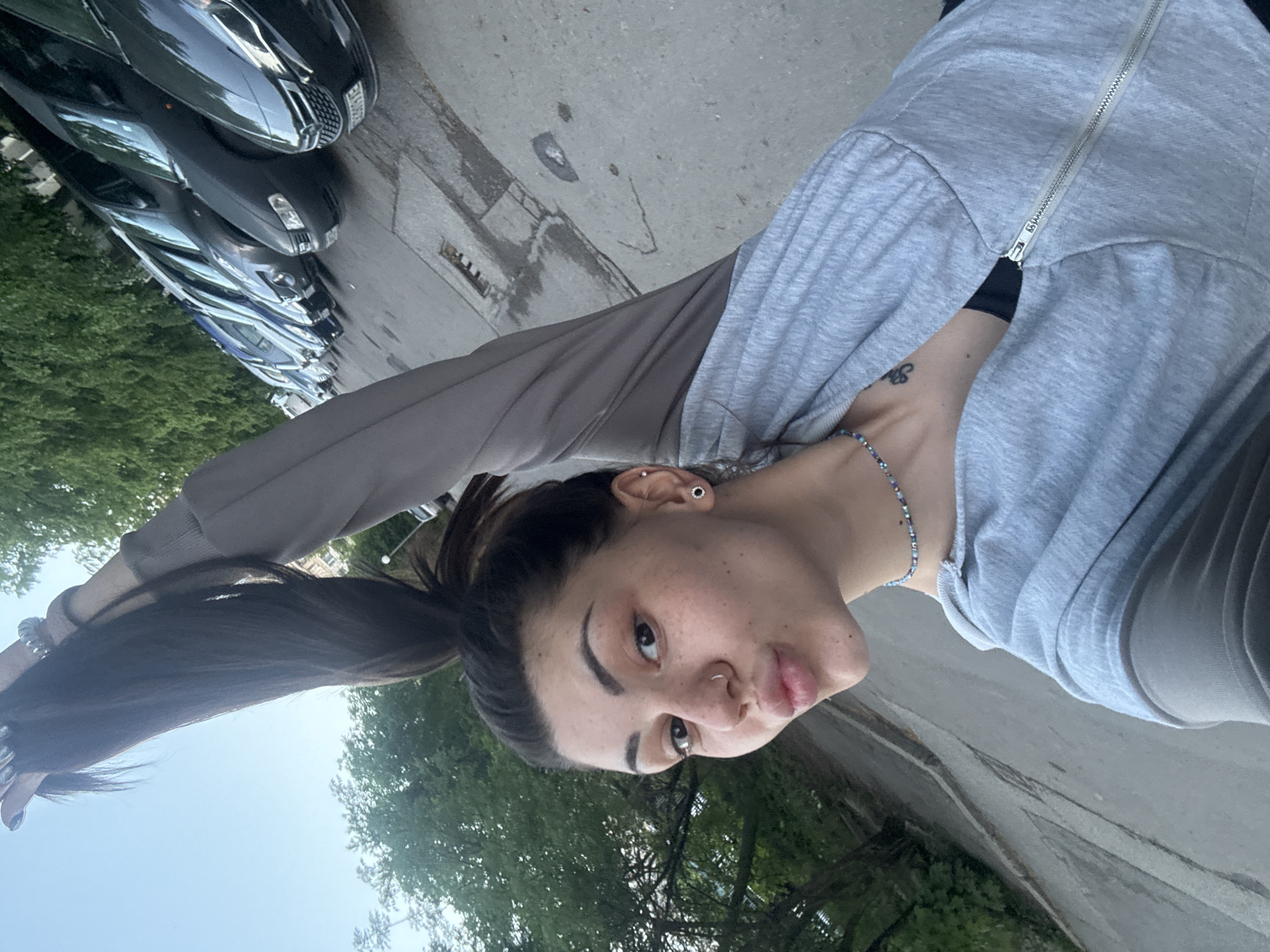 A young woman is taking a selfie outdoors. She is fair-skinned with dark hair tied in a high ponytail, and is wearing a grey zip-up hoodie over a dark top. She is wearing a bracelet on her wrist and a necklace. Her hair is held by a silver band. She is puckering her lips slightly for the photo.
In the background, there are trees, a sidewalk, and parked cars. The location is plausibly in the city of Sofia, Bulgaria, given the types of cars and the architecture, which is visible between the trees.