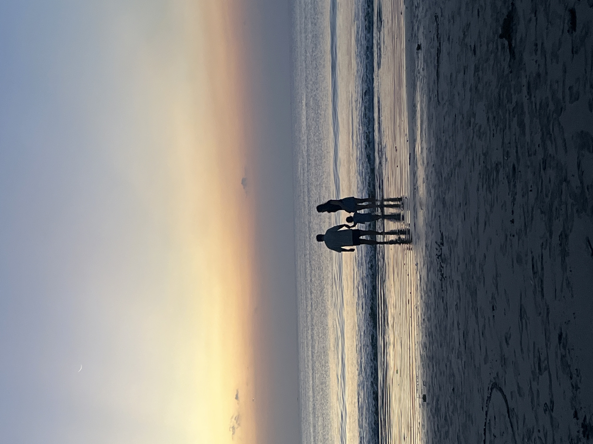 In Kuta Selatan, Indonesia, at sunset, a family of three silhouetted figures stands near the ocean's edge on a sandy beach. The parents stand side-by-side, with a small child between them, all facing the water. The sky displays a gradient of colors, transitioning from a pale yellow near the horizon to a soft blue at the top. A sliver of a crescent moon is visible in the upper left portion of the sky. The ocean is calm, with gentle waves lapping at the shore, reflecting the colors of the sunset. The wet sand in the foreground creates a mirror-like surface, reflecting the silhouettes of the family and the sky.