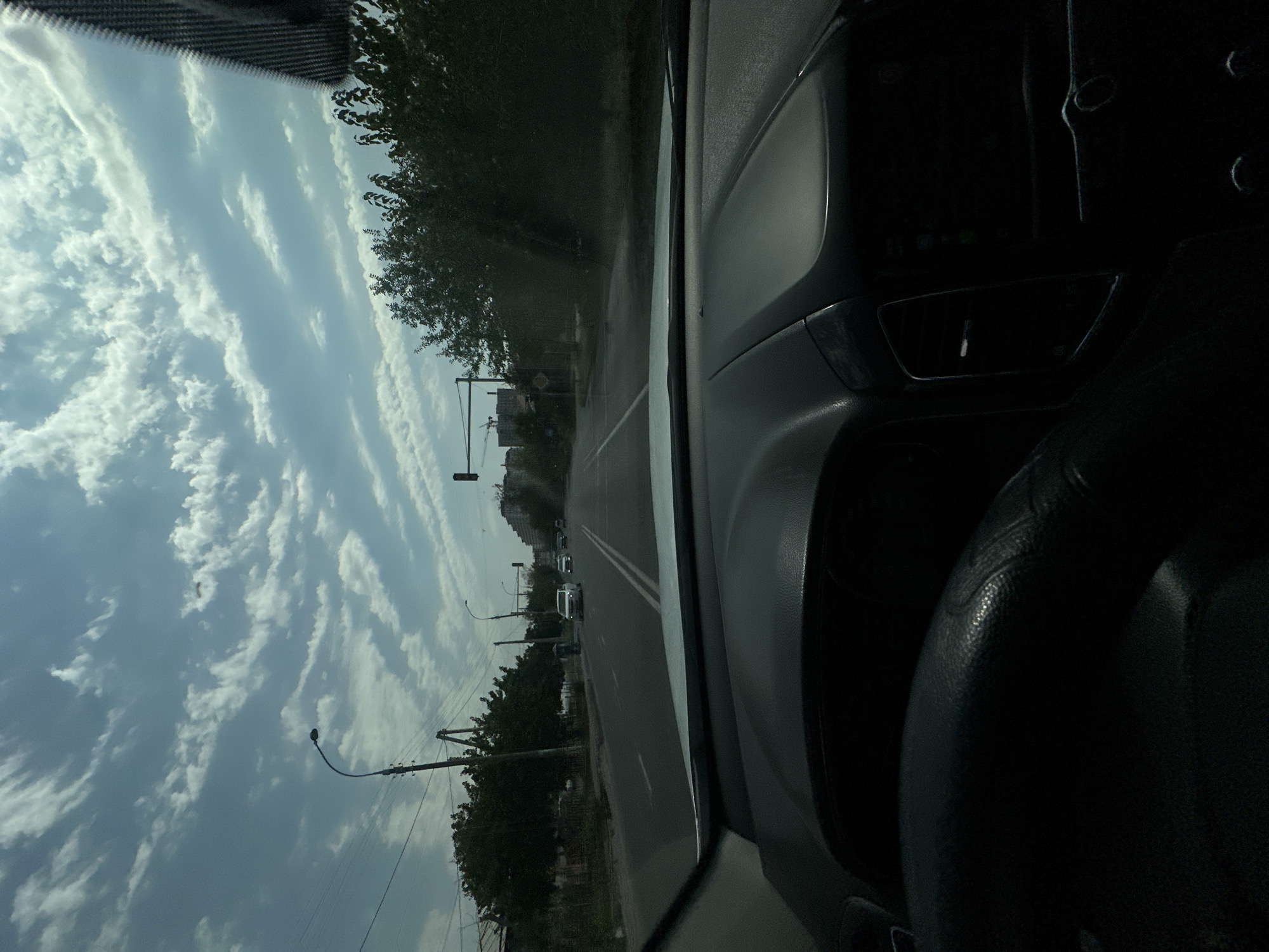 The image is taken from the interior of a vehicle looking forward through the windshield. The view is of a wide, paved road in Tashkent, Uzbekistan, lined with trees and utility poles. The road has multiple lanes and appears to be in an urban or suburban setting. In the distance, buildings are visible. The sky is partially cloudy with scattered white clouds against a blue background. There are several other vehicles on the road, including cars and what appears to be a white minivan. A traffic light is visible in the distance. The interior of the car is dark, with the dashboard and steering wheel partially visible in the foreground.