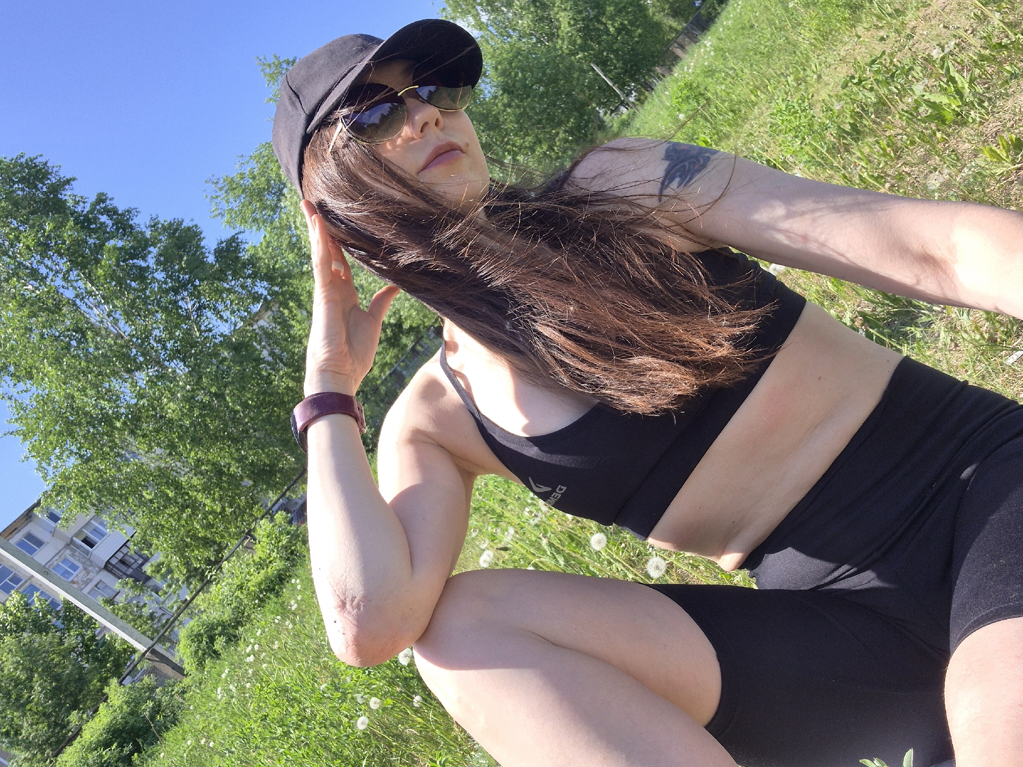 A woman is squatting in a grassy area. She is wearing a black sports bra, black shorts, a black cap, and sunglasses. A tattoo is visible on her left upper arm. She is wearing a purple fitness tracker on her left wrist and has her left hand near her head. The grass is green and interspersed with dandelions. Behind her, there are green trees and a light-colored apartment building with windows. The sky is blue.