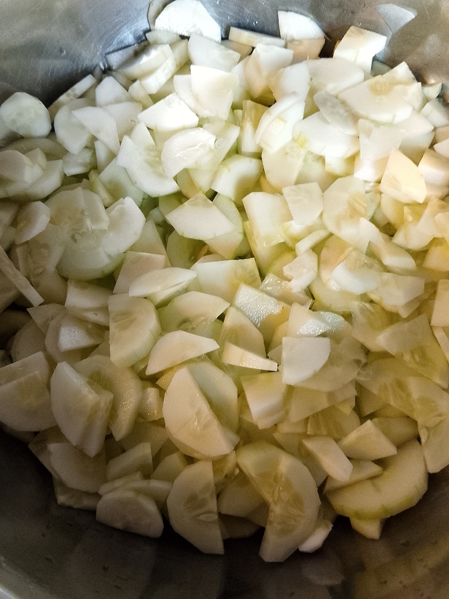 A close-up, overhead shot shows a metal bowl filled with chopped cucumbers. The cucumbers are sliced into thin, irregular pieces, some appearing to be half-moons and others more angular chunks. The light source from above creates highlights and shadows, giving the translucent cucumber slices depth. The bowl itself is a reflective, brushed metal. The image is focused on the contents of the bowl, with the edges of the metal container visible at the top and bottom. There is no visible indication of location in Lhokseumawe, Indonesia.