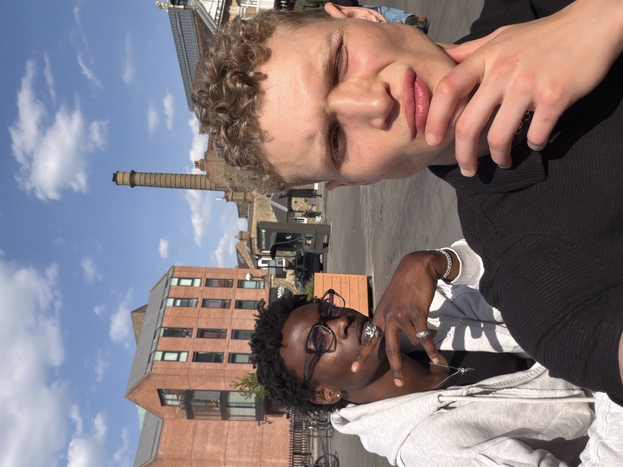 This is an outdoor, vertical selfie-style photograph featuring two individuals in an urban setting, likely in London, United Kingdom. The image is taken during the daytime under a bright blue sky with scattered white clouds.
The subject in the foreground, positioned prominently on the right side of the frame, is a person with light skin, short curly brown hair, and light eyes, partially squinting. They are wearing a dark top, and one hand, with fingers curled, rests near their mouth and chin. Their expression appears casual and relaxed, possibly squinting from the sunlight.
Behind and to the left of the first person is a second individual with dark skin, dark curly hair, and glasses. This person is wearing a light grey hooded sweatshirt and various silver accessories, including multiple rings on their fingers, a silver bracelet on their left wrist, and a silver necklace with a small pendant. They are looking slightly away from the camera, posing casually.
The background reveals a bustling city environment. Several buildings constructed from brick and other materials are visible. A tall, slender brick chimney or smokestack stands prominently against the sky in the mid-ground. The ground appears to be a paved street or public square. Some blurred vehicles and other street elements are also discernible in the distance. On a black sign in the background, partially readable text includes "APPELLAVO" and some other indistinct words. Another small sign nearby might show "50" or "SO".