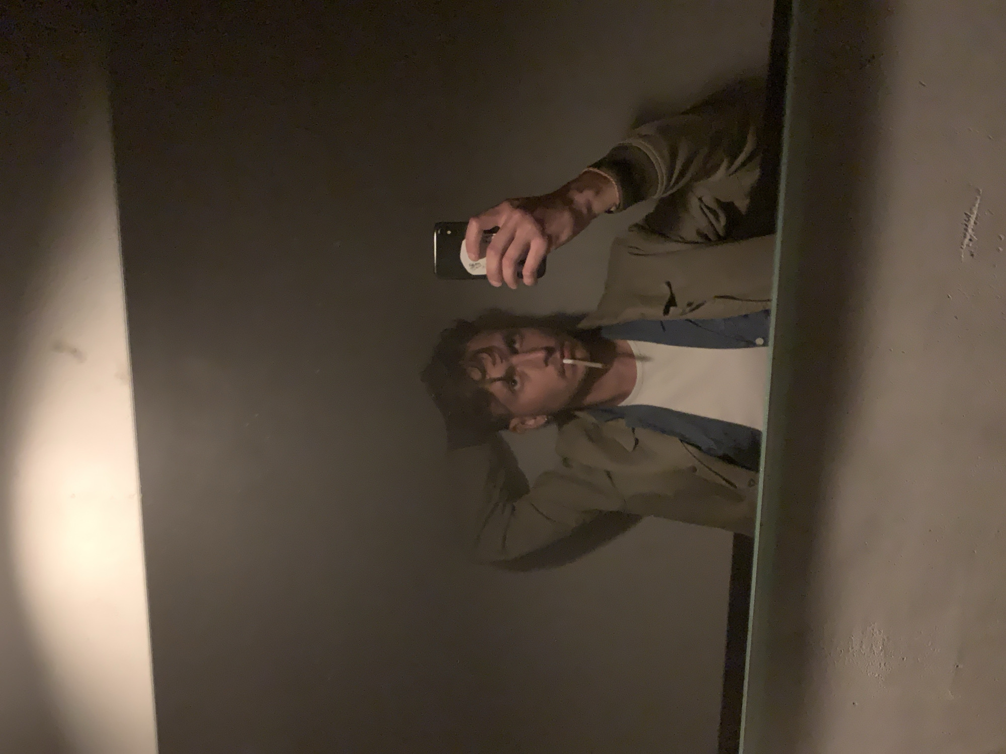 A mirrored selfie shows a man holding a phone. The man, with curly hair, has a cigarette in his mouth and is wearing a white shirt with a blue denim shirt. The mirror is in an enclosed space with walls of a muted gray color. There is no visual information in the image to determine if the location is in Tel-Aviv, Israel.