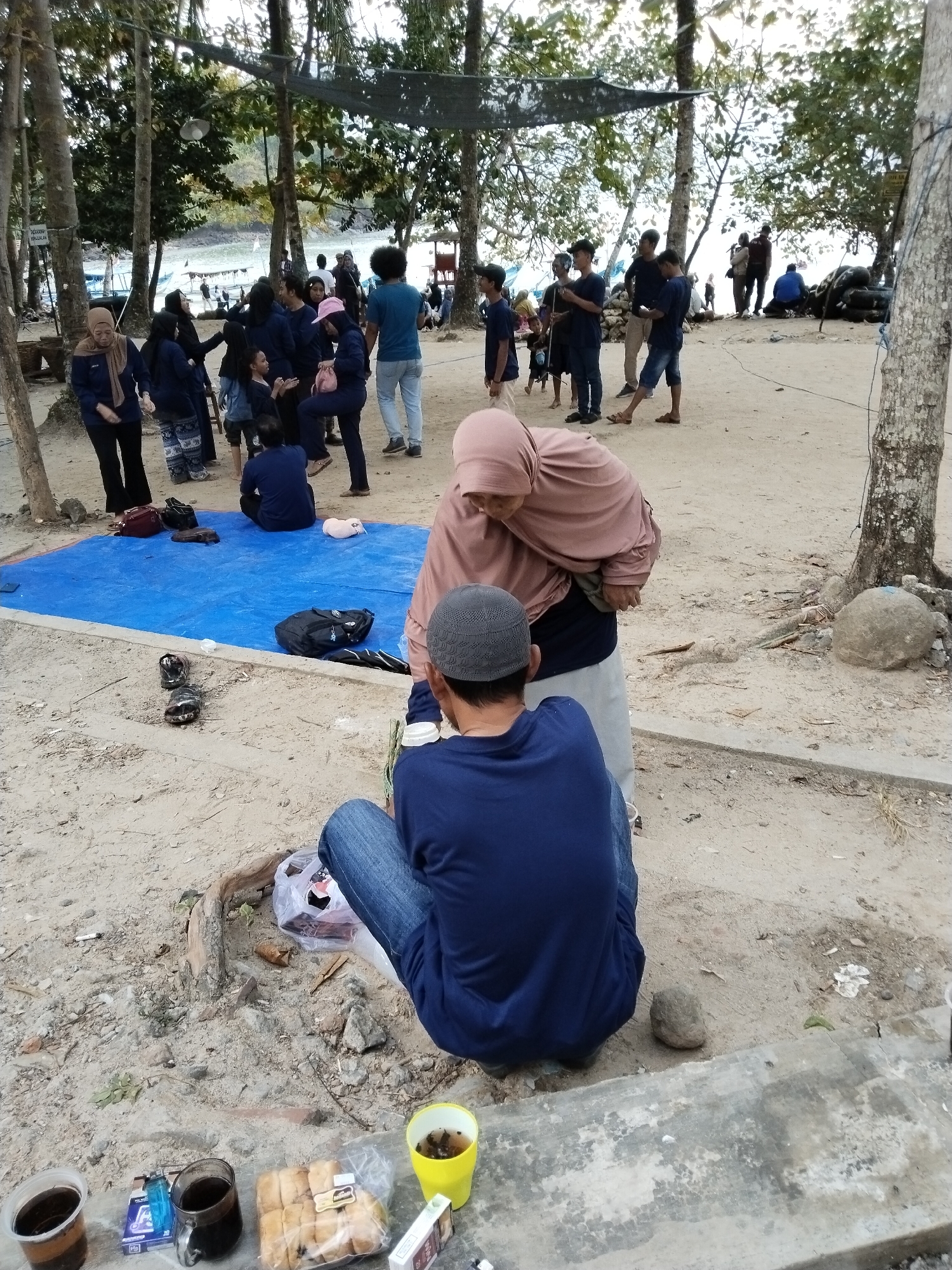 In Prigi, Indonesia, on a sandy beach area under scattered trees, a group of people are gathered. In the foreground, a man wearing a blue t-shirt and jeans, and a grey skullcap sits on the ground facing away from the camera. To his left, a woman in a light pink hijab and dark clothing leans over, also facing away from the camera. Scattered around them are various items: two cups of dark liquid, a pack of cigarettes, a lighter, a plastic bag containing bread rolls, and a yellow cup with a beverage. A blue tarp is spread on the sand to the left, with bags and other items on it. In the background, a larger group of people, many wearing navy blue shirts, are visible standing and interacting near the water's edge. The environment suggests a casual outdoor gathering, possibly a picnic or break during an activity.