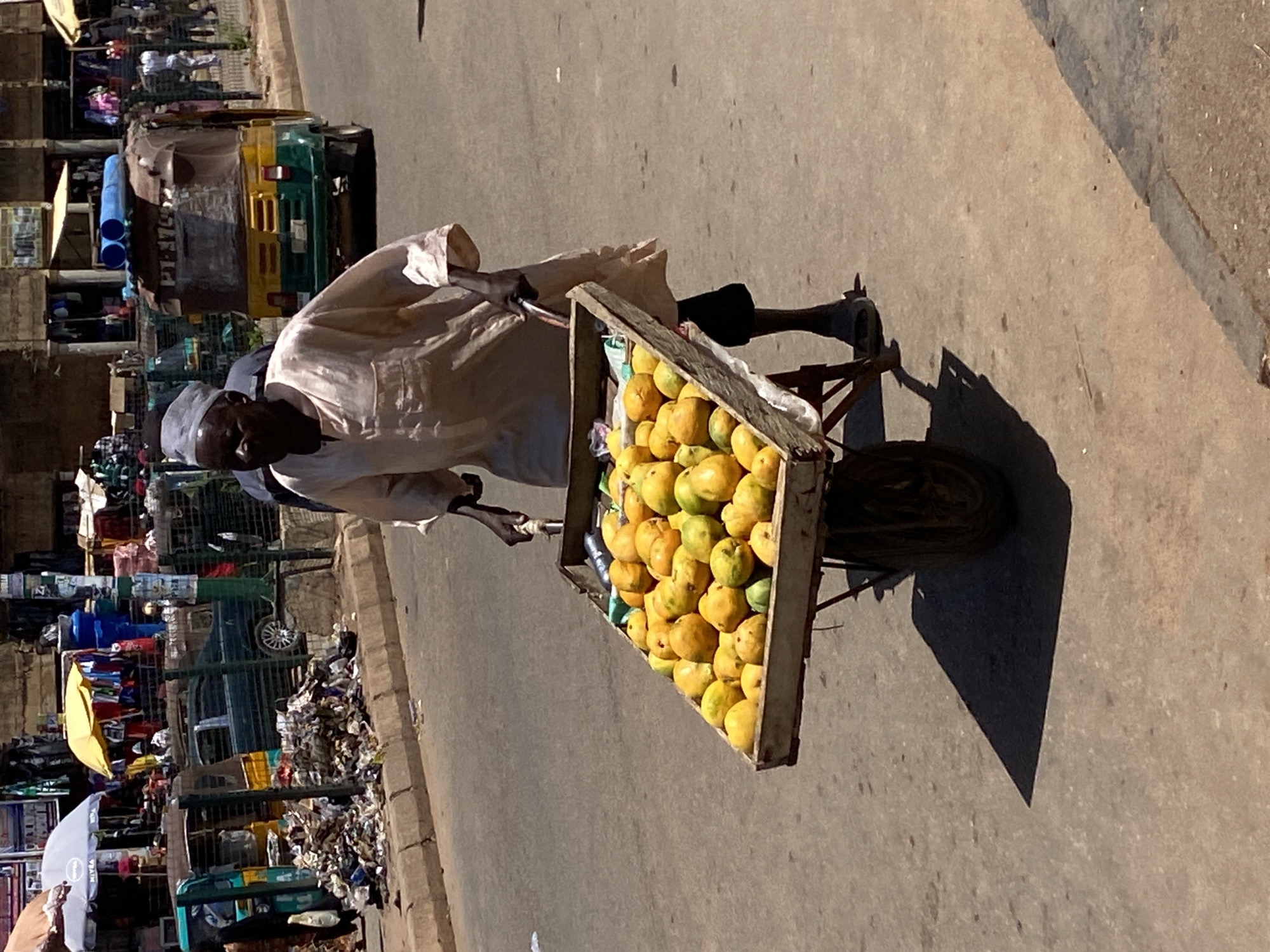 The image shows a man in Bukuru, Nigeria, pushing a wheelbarrow filled with mangoes. He is wearing a light-colored robe and a head covering. The wheelbarrow is wooden, and the mangoes are piled high. The street is paved and lined with shops and vehicles, including a three-wheeled taxi or "keke." There is also a substantial amount of trash visible along the side of the road. The scene is bathed in sunlight.