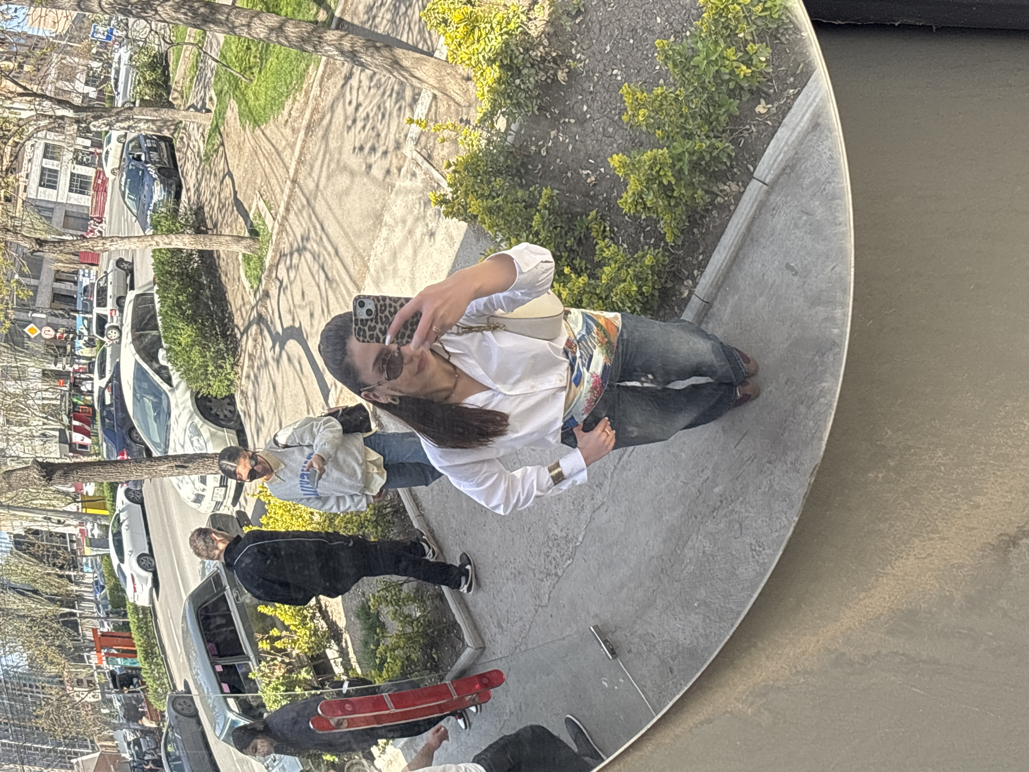 The image, rotated approximately 90 degrees counter-clockwise, presents a reflection in a large, circular convex mirror, capturing an urban street scene in Yerevan, Armenia, on a sunny day.
In the foreground, a woman is seen taking a selfie in the mirror with a smartphone encased in leopard print. She wears sunglasses, a white button-up shirt, blue jeans, and a colorful patterned scarf or belt, with a small white shoulder bag. Her long dark hair flows over her shoulder. Next to her, another person in a light grey sweatshirt with partial text "UNI" visible, sunglasses, and jeans, also appears to be looking towards the mirror or taking a photo. Further left, a person in a black jacket and pants is seen from behind, walking or standing. A fourth person, partly visible in the lower right, is near a red skateboard with white wheels.
The background, reflected around the people, depicts a city street lined with trees and various parked cars, including white, blue, and dark grey models. Some trees have green foliage, while others appear more bare, suggesting spring or early autumn. Sidewalks and bushes are visible, along with buildings further in the distance. The bright sunlight casts strong shadows, indicating a clear, sunny day. The scene suggests a casual outdoor setting, possibly a public square or boulevard, where people are enjoying the weather and interacting with the reflective surface.