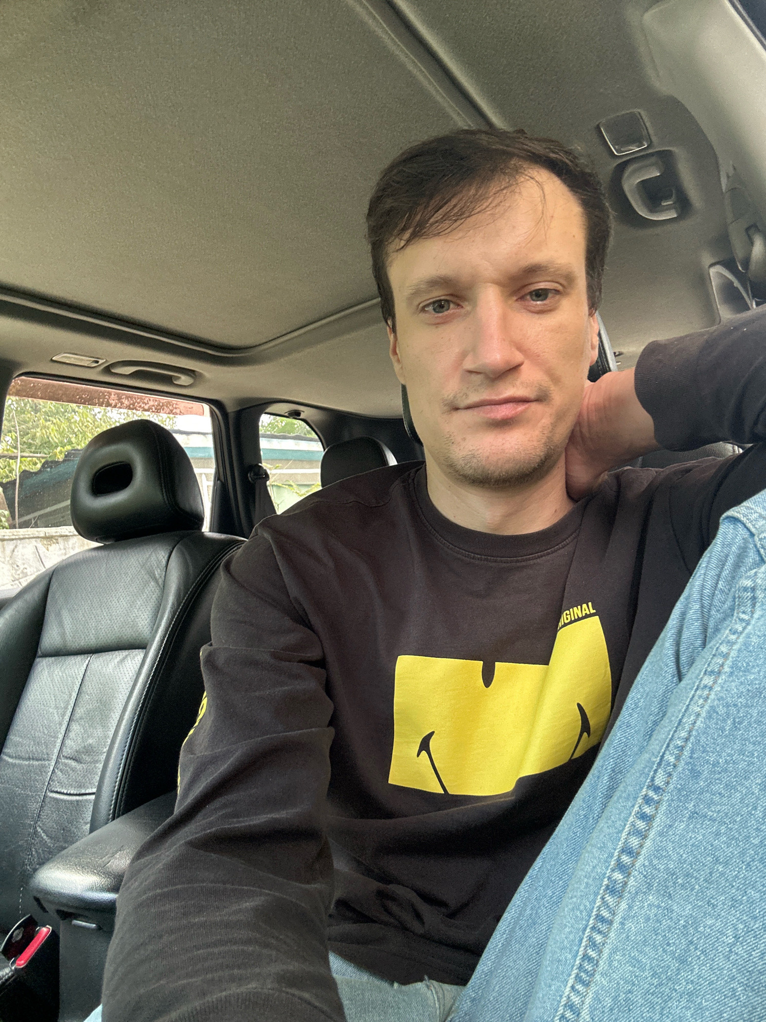 In Makiv, Ukraine, a man is seated in the driver's seat of a vehicle, captured in a selfie-style photograph. The car's interior is visible, featuring black leather seats, a sunroof, and interior lighting. The man, with medium brown hair and fair skin, wears a dark long-sleeved shirt adorned with a large yellow smiley face graphic and the word "ORIGINAL" in yellow lettering. He is also wearing light blue denim jeans. His left arm is resting behind his head, and he is looking directly at the camera with a neutral expression. Outside the car windows, trees and some buildings can be seen, suggesting an outdoor, possibly suburban or rural, setting.