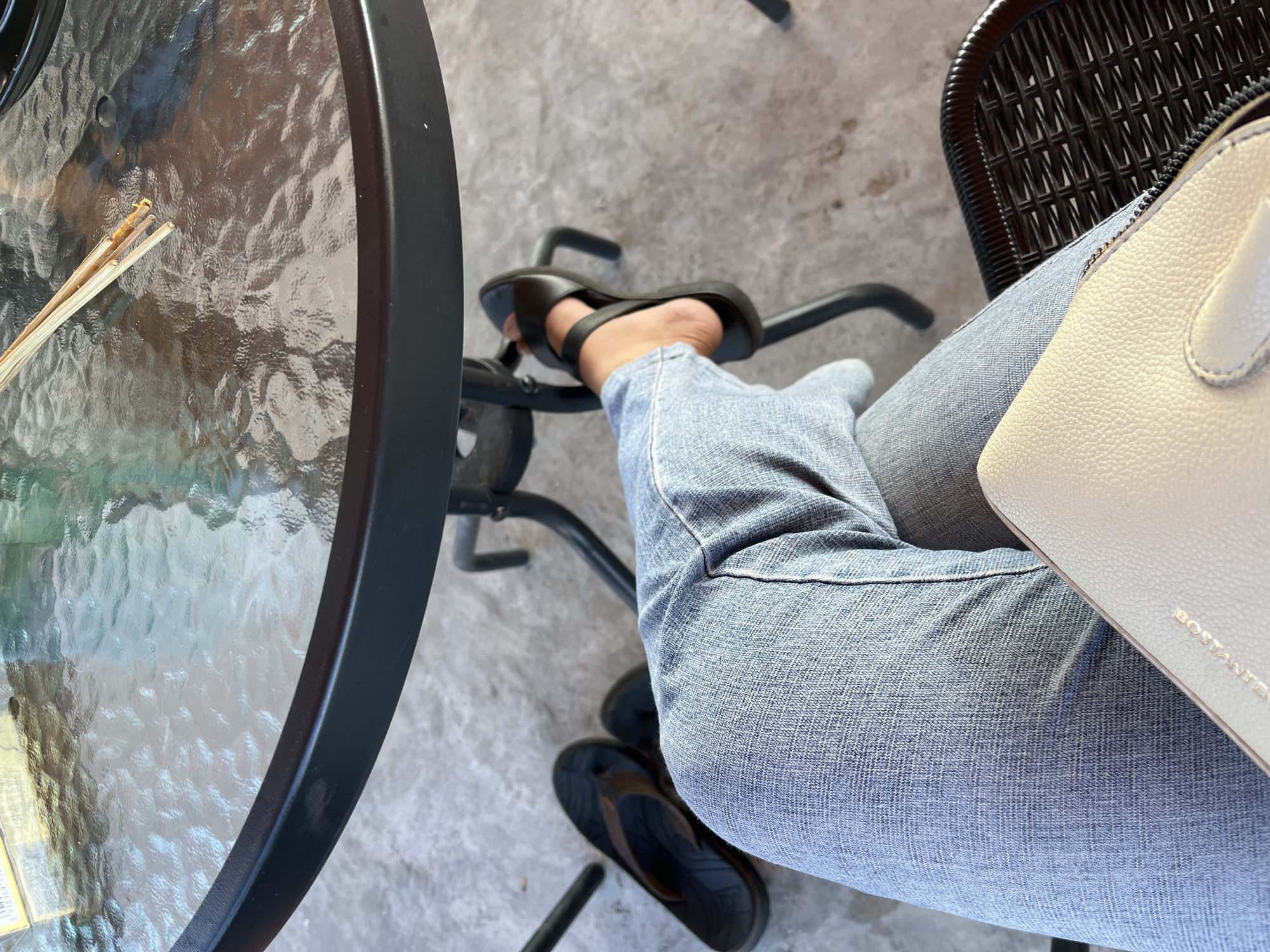 A person sits at an outdoor glass-topped table in Oro-oro Ombo, Indonesia. They are wearing light blue jeans and black sandals. Their left foot is resting on the leg of a black metal table. A pair of black flip-flops are on the ground near the table. A light-colored purse is on the person's lap. Two wooden chopsticks are on the table, with some food remnants on them. The ground appears to be concrete.