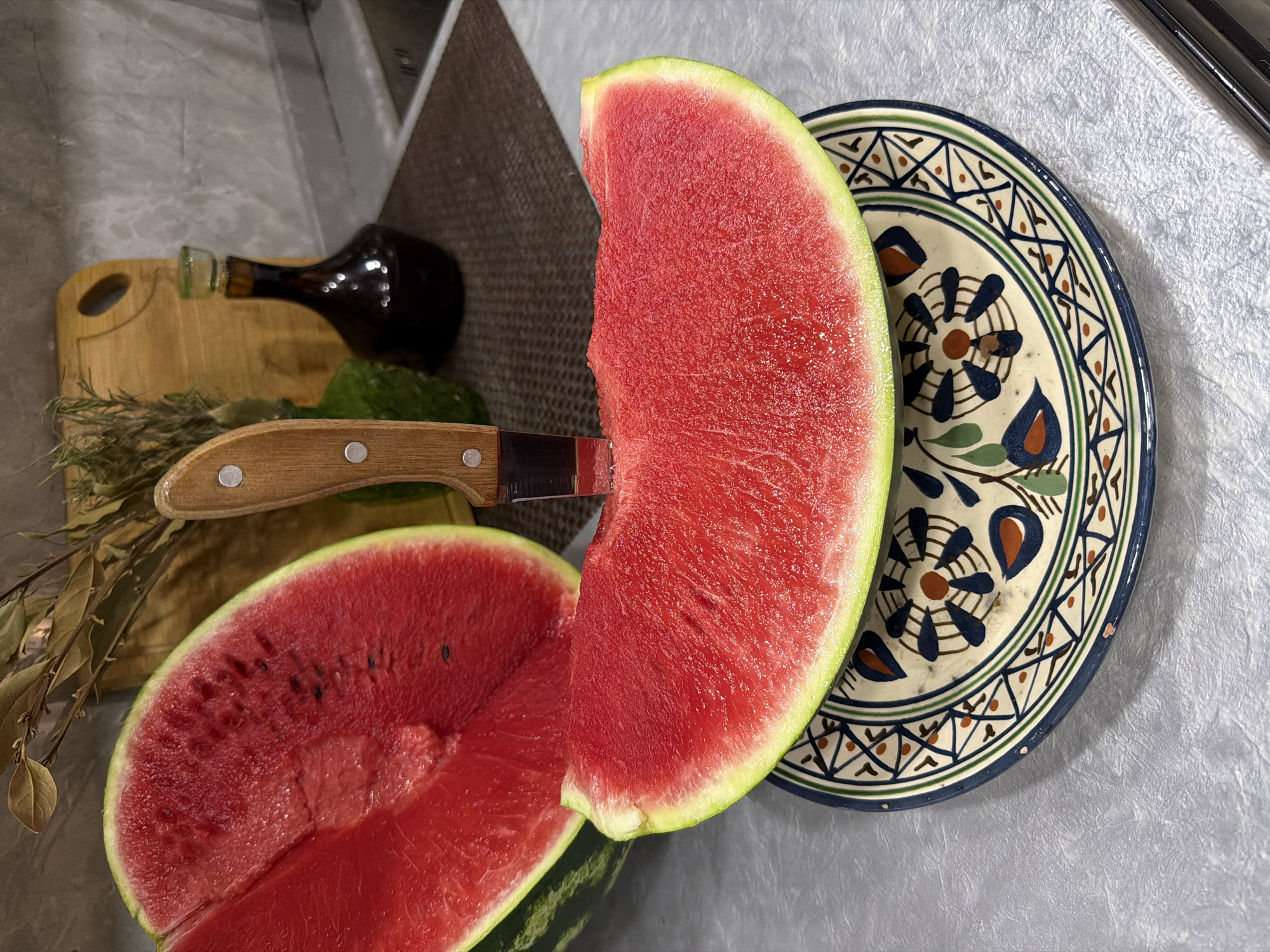 An overhead shot captures a kitchen scene in Sabirabad, Azerbaijan, featuring a slice of watermelon resting on a decorative plate. The watermelon slice, with its vibrant red flesh and light green rind, is positioned prominently in the foreground. A knife with a wooden handle is embedded in the watermelon. To the left, a halved watermelon is visible, revealing its juicy interior and dark seeds. In the background, a wooden cutting board, a dark glass bottle, and dried plant sprigs add to the kitchen setting. The countertop surface appears textured, and the overall lighting suggests an indoor environment. There are no people present in the image.