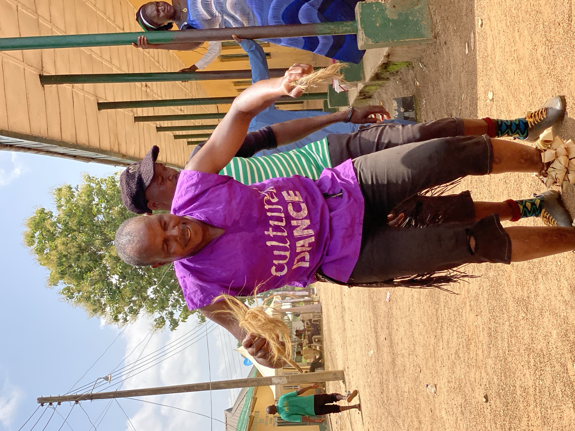 A street scene in Maigizo, Nigeria, depicts a smiling person wearing a purple shirt with the words "culture DANCE" in white text, and dark shorts. They are holding a bundle of straw-like material in one hand and have bundles tied around their lower legs. A person with a hat and a striped shirt is partially visible behind the person in the purple shirt. Others are visible in the background, including people on a raised structure and a person in a green shirt walking along the ground. A utility pole and building can also be seen, with trees in the distance and a sandy ground surface.