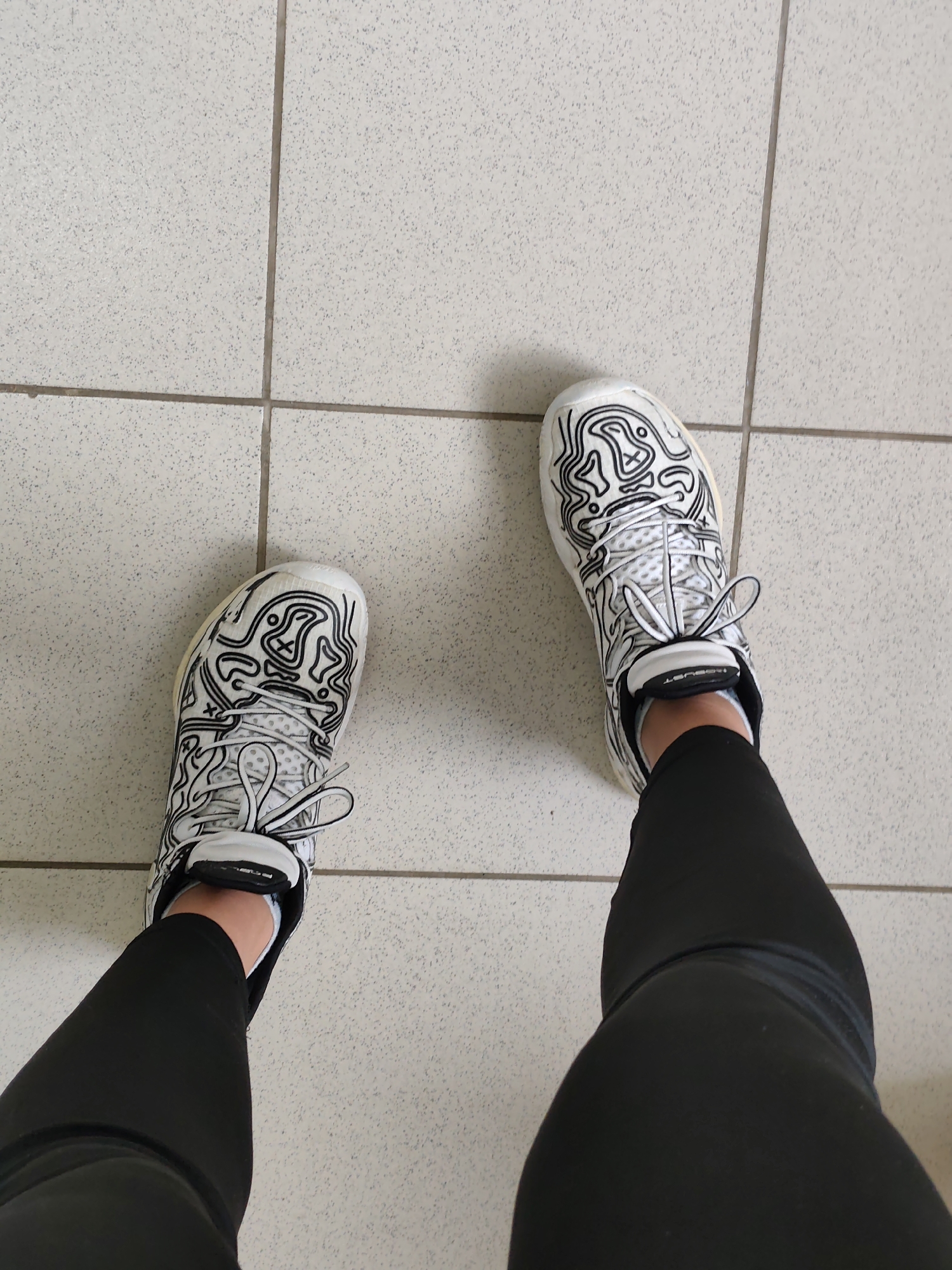 An overhead shot shows a pair of white sneakers with black abstract line patterns and black leggings on a person's legs. The sneakers have white laces and black logos on the tongue. The person is standing on a tiled floor with a light grey speckled pattern and visible grout lines. The location in the city of Chaikovsky, Russia, is not discernible from the image.