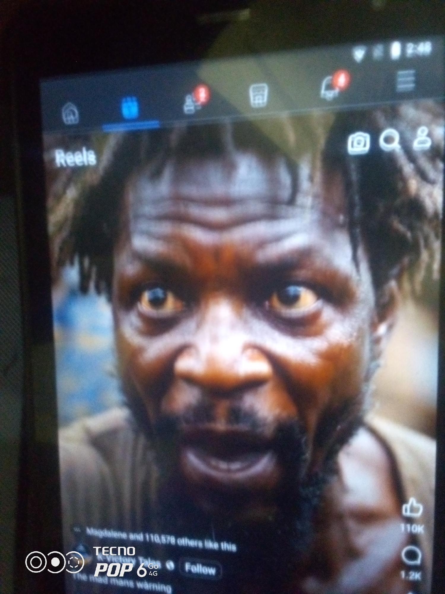 The image is a close-up, slightly blurry shot of a man's face displayed on a mobile device, likely a smartphone. The man has dark skin, short, messy dark hair, and his eyes are open with a yellowish tint. His expression appears intense or distressed. The screen shows a social media interface, with the word "Reels" visible in the upper left corner. Other icons and text, including "Tecno Pop 6 4G" and "Follow," are also present. The background of the video appears to be a blurred outdoor scene with blues and earthy tones. There is no information about the location within the city of Howayi, Nigeria, or any other geographical context. The image captures a moment from a digital video being viewed on a phone.