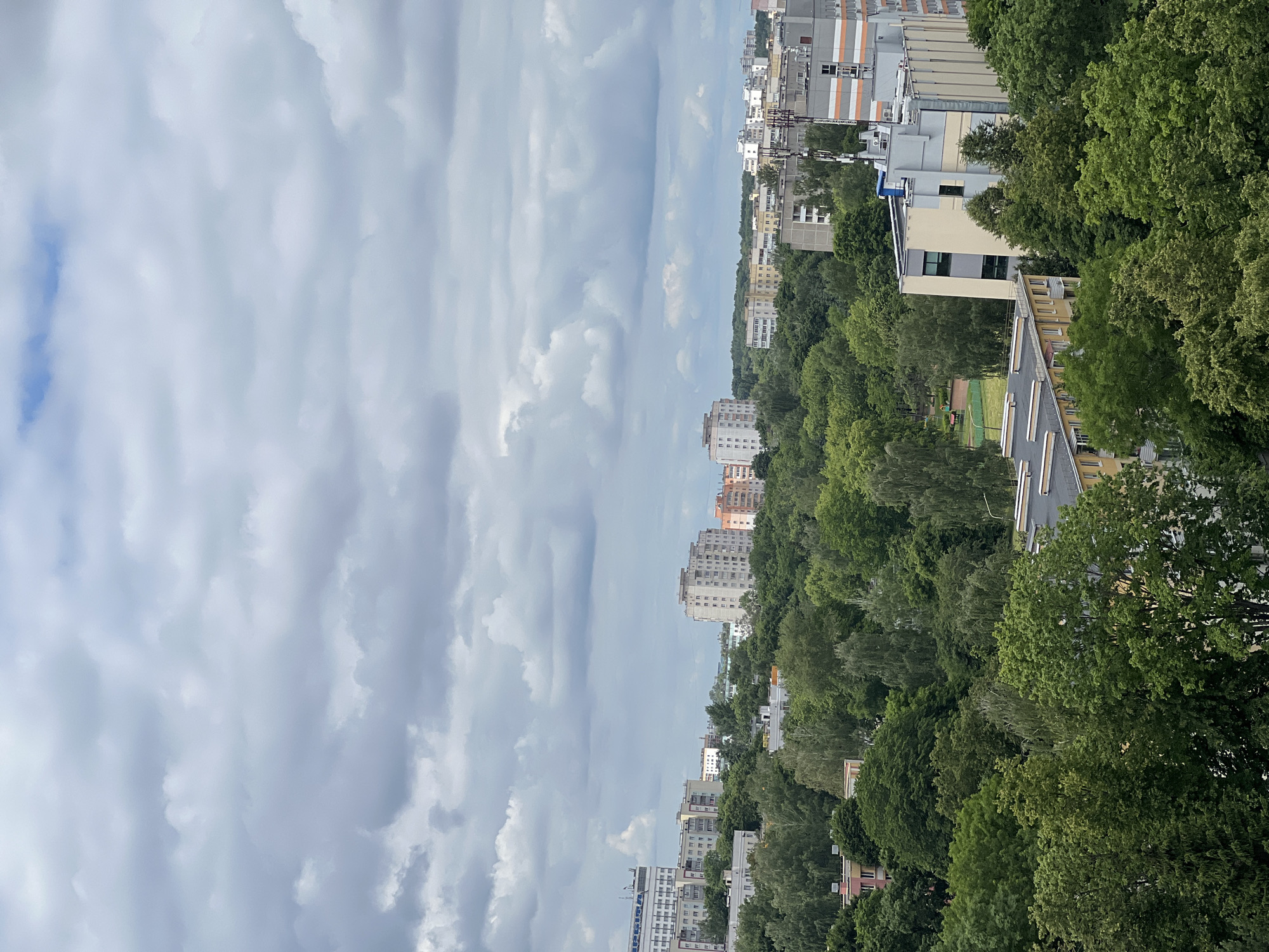 The image is an aerial view of a cityscape in Minsk, Belarus. It features a dense canopy of green trees interspersed with various buildings, ranging from apartment complexes to more modern structures. The sky is overcast with a thick layer of gray clouds, with hints of blue peeking through in some areas. In the foreground, a cluster of trees partially obscures a yellow apartment building with a dark roof. Further back, a park area with green fields and playground equipment is visible, bordered by more trees. The middle ground is dominated by a lush forest that separates the foreground from a line of apartment buildings in the background. These buildings vary in height and architectural style, with some featuring balconies and multi-colored facades. In the distance, taller buildings rise above the tree line. A telecommunications tower is prominent on the right side of the image, alongside a multi-story building with orange and gray detailing. The overall impression is one of a city that has integrated nature into its urban fabric, with significant green spaces within its built environment.
