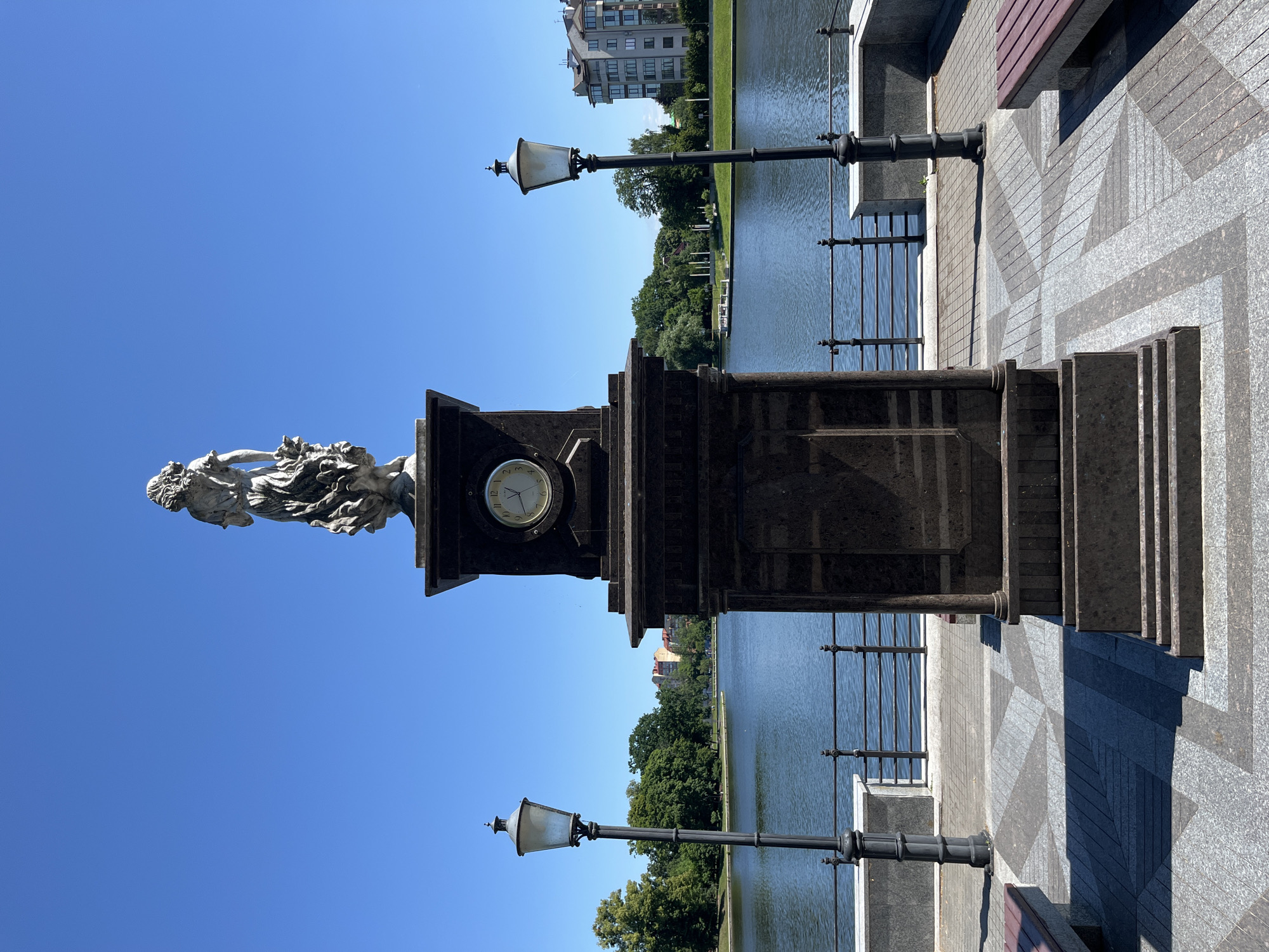 This vertical, eye-level photograph captures a scenic waterside promenade on a bright, clear day. The primary subject is a stone monument featuring a clock face and a sculpted female figure, likely a statue. The monument is made of dark granite and has a classical design. A lamppost with a white lantern is positioned to the left of the monument.
The setting appears to be a public park or waterfront area, with a body of water, possibly a canal or river, to the right of the monument. Across the water, a lush green treeline separates it from a row of multi-story buildings in the distance, suggesting an urban environment. The promenade itself is paved with granite tiles in a patterned design, with a darker granite base supporting the monument. A wooden bench with a red seat is partially visible in the foreground. Another lamppost, similar to the first, stands further down the promenade.
The bright sunlight and clear blue sky indicate it is daytime, likely mid-morning or early afternoon, with fair weather. No people are visible in the frame, giving the scene a tranquil and serene atmosphere. The image's orientation is rotated 90 degrees counter-clockwise, so the water and buildings appear to be on the left side of the frame, and the sky is at the top. There is no visible text in the image.