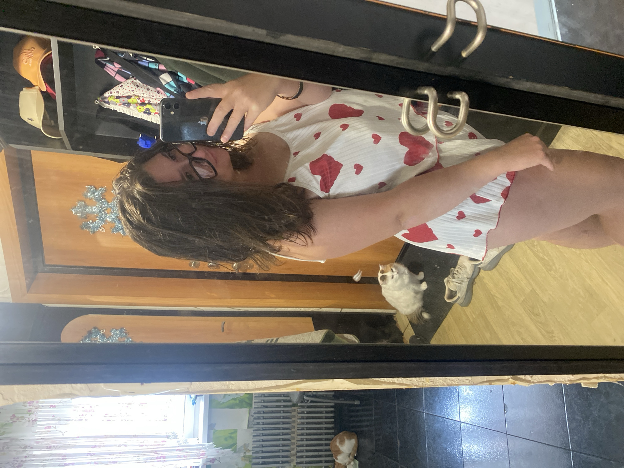 In a residential interior, a young woman with dark, wavy hair and glasses poses for a selfie in a full-length mirror. She is wearing a white pajama set with red hearts. The reflection reveals a room with wooden doors, a window with curtains, and various items on shelves, including hats and clothing. In the background of the reflection, two cats are visible on the floor, one orange and white, and the other white with orange markings. The scene suggests a casual, domestic setting, possibly within a home in Chaikovsky, Russia.