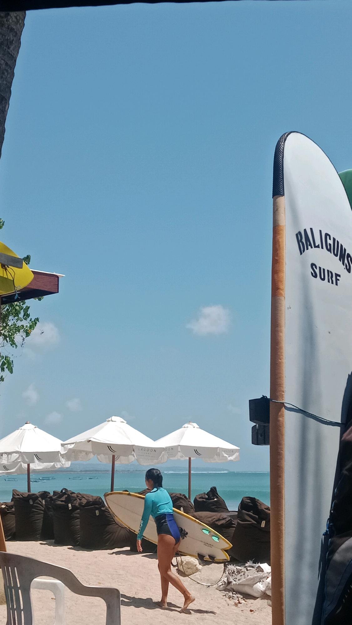 A person, possibly a woman, wearing a turquoise and black wetsuit is walking on a sandy beach in Kuta, Indonesia, carrying a white surfboard with yellow accents. The surfboard is pointed towards the right side of the frame, and the person is facing away from the viewer, with their left leg extended forward. Behind the person, there are several white beach umbrellas set up on the sand. Further back, a body of water, likely the ocean, is visible with gentle waves. In the foreground, a portion of a white chair is visible on the left side. To the right of the surfboard carrier, a tall, white surfboard with black lettering that reads "BALIGUNS SURF" is standing upright. The sky is clear and blue with a few small clouds. The overall scene depicts a typical beach setting in Kuta.