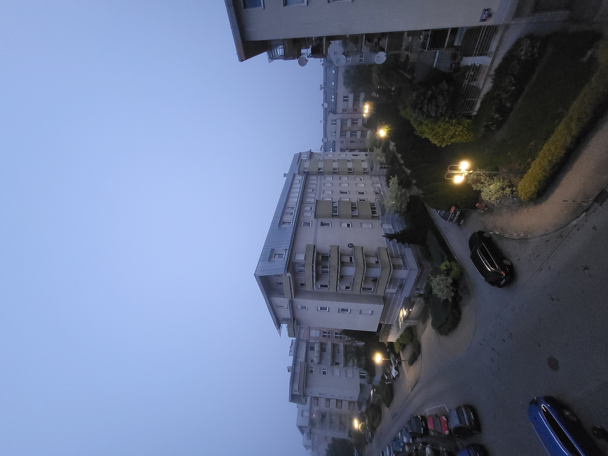 The image presents an elevated view of a quiet residential area at what appears to be dawn or dusk, characterized by soft, dim natural light and illuminated streetlights. A vast expanse of pale blue-grey sky, suggesting misty or hazy conditions, fills the upper and left portions of the frame.
In the middle and right, several multi-story apartment buildings are visible, featuring uniform windows and balconies. One prominent building displays multiple satellite dishes attached to its balconies. The architecture suggests modern residential structures, predominantly in shades of light grey, beige, and white.
A winding, paved road or driveway snakes through the scene, flanked by sidewalks and sections of green landscaping including bushes, hedges, and small trees. Numerous cars are parked along the road and in parking areas, including a distinct black car and a partially visible blue car. Warm yellow light emanates from several streetlights, casting glows on the ground and foliage. No people are visible, contributing to a tranquil and still atmosphere. A small, indistinct sign, possibly a building number, is barely legible on one of the buildings.