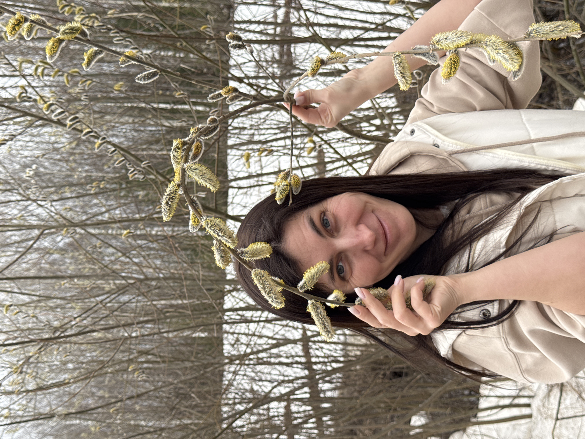 A woman with long dark hair and blue eyes is holding a pussy willow branch in her hands, appearing to be outdoors. She's wearing a light beige jacket with a white puffy vest underneath. The background consists of many bare tree branches, suggesting it might be late winter or early spring. The location seems to be in a forest or wooded area.
