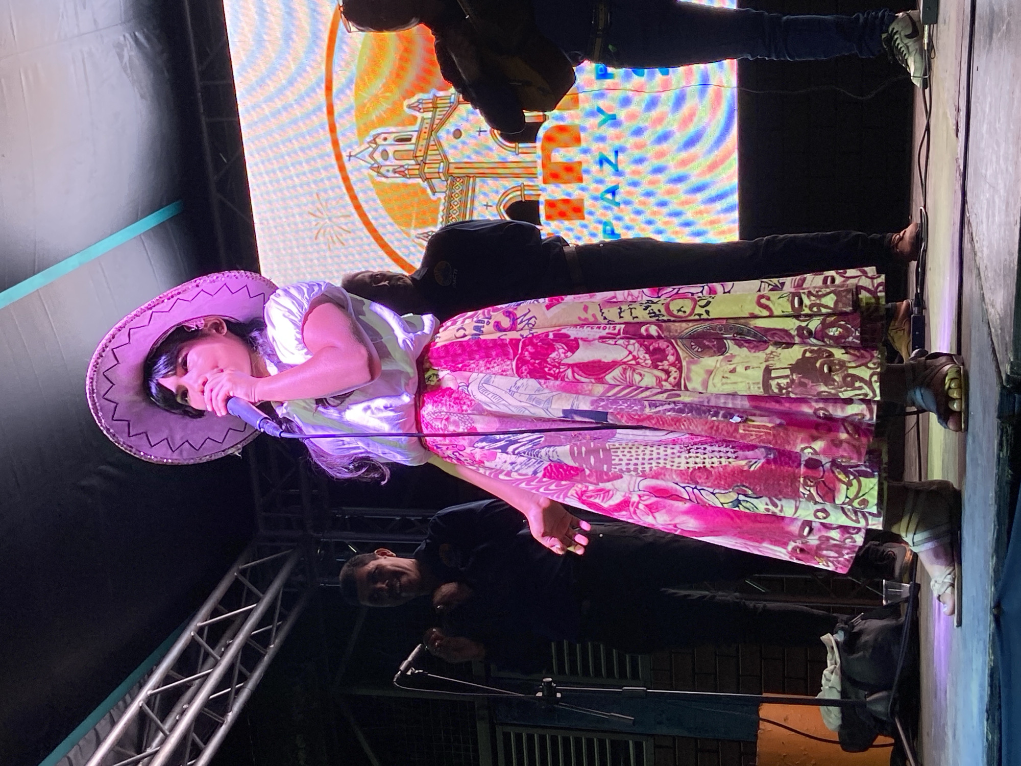 The image captures a scene from Tinaquillo, Venezuela. A female singer is at the forefront, holding a microphone and wearing a light-colored cowboy hat, a white shirt, and a long, vibrant patterned skirt with pink and yellow hues. Behind her, several individuals appear to be part of a band or performance group. They are dressed in dark clothing. In the background, a large screen displays the word "Tinaquillo" vertically beside an illustration of a building. The setting appears to be an outdoor stage, illuminated by stage lights. Metal scaffolding is visible to the left of the singer. The overall composition suggests a cultural event or performance taking place in the city.