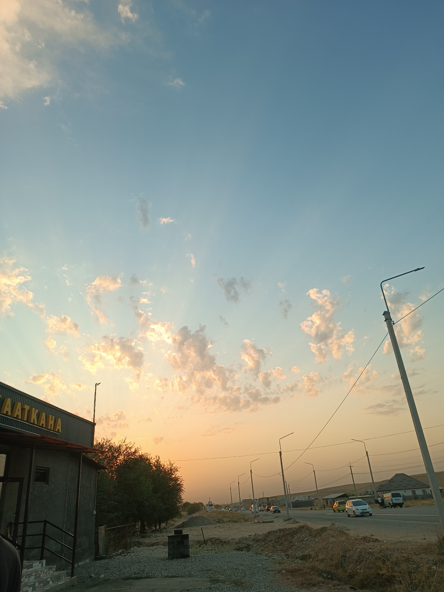 The image depicts a view along a road in the city of Jangy-Akman, Kyrgyzstan, during what appears to be sunset. The sky is a gradient of light blue and orange, with scattered clouds illuminated by the setting sun.
On the left side of the frame, there is a building with the word "AATKAHА" visible in yellow letters on its facade. Adjacent to the building is a covered entryway with stairs leading up. Several trees line the area behind the building.
The road itself is visible in the mid-ground and extends into the distance. Several vehicles are driving on the road, including cars and what appears to be a van. Streetlights are present along the roadside. In the background, there are houses and other structures, suggesting a semi-urban or rural setting. The ground in the foreground is covered with gravel and dry vegetation, and there is a pile of dirt and rocks. A lamppost is prominently featured on the right side of the image. The overall scene suggests a somewhat arid environment.
