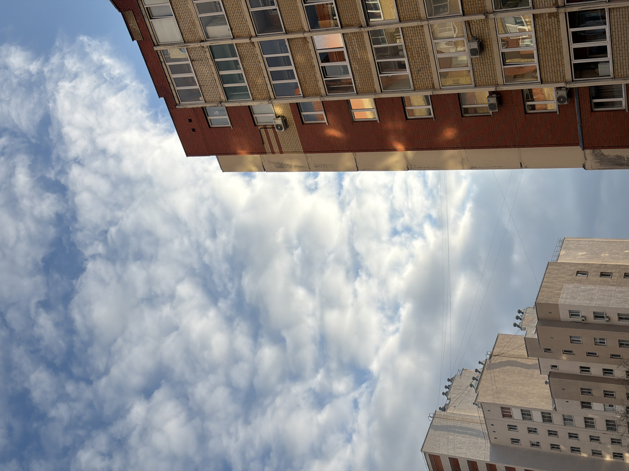The image presents an upward-angled view of the sky and the architectural details of two buildings in Penza, Russia, under a partly cloudy daytime sky. The perspective, possibly due to image rotation, positions the buildings diagonally opposite each other.
The building occupying the upper left quadrant features a lower section of reddish-brown brick, topped with lighter-colored walls. It is characterized by multiple rows of rectangular windows, some reflecting the bright sky and warm sunlight, while others reveal interior glimpses. Several air conditioning units are visible on its facade.
In the lower right, another building stands, distinguished by its light beige or grey exterior and a stepped, terraced design. This structure also has numerous windows and some visible external vents or smaller air conditioning units. A network of thin wires or cables spans across the mid-ground sky, appearing to connect to or pass by this building.
The central and largest portion of the frame is filled with a blue sky dotted with white and grey cumulus clouds, indicating a partly cloudy day. The sun's presence is evident from the reflections and highlights on the building windows. There is no discernible human activity or visible text within the frame.