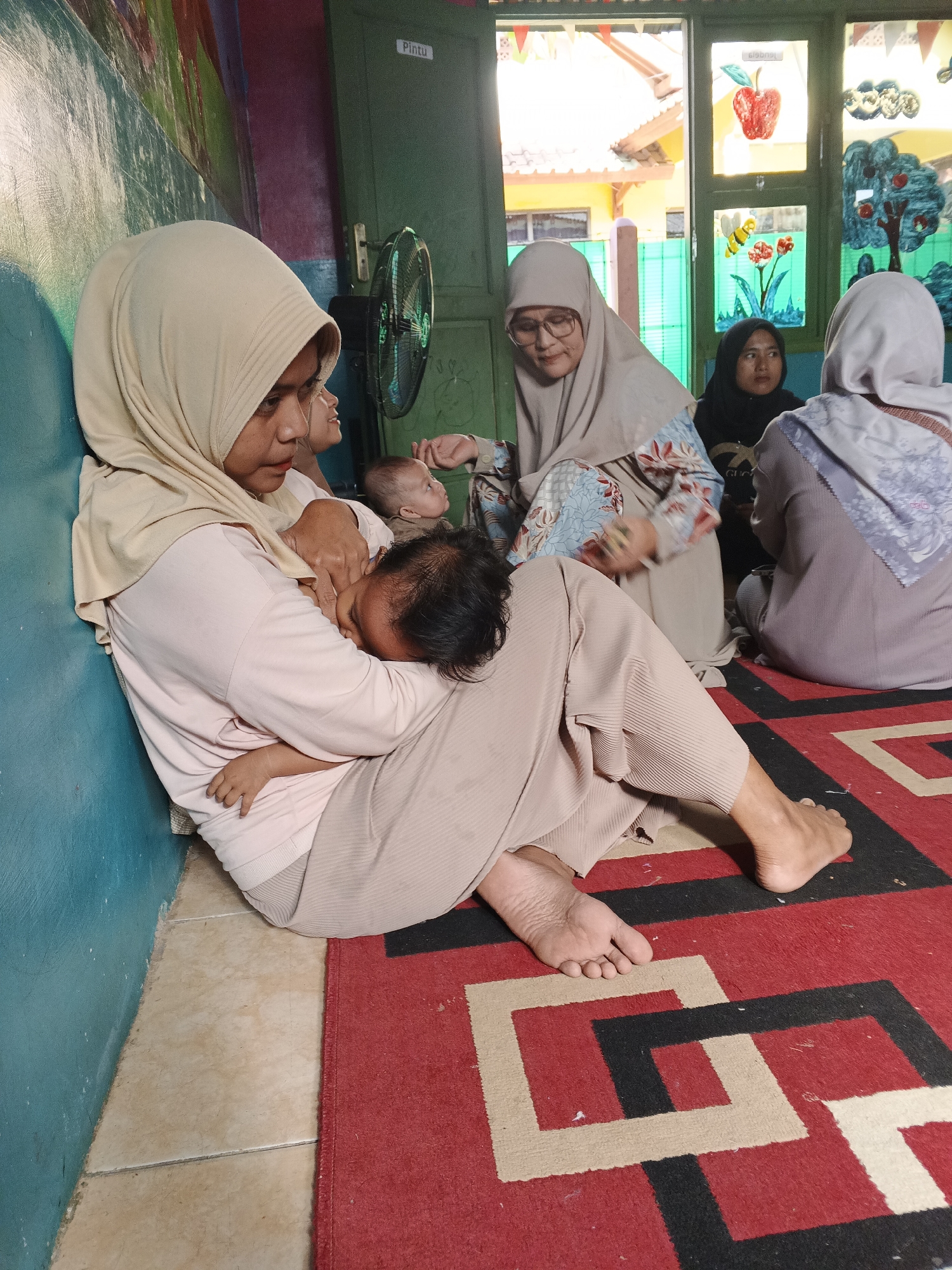 A woman, wearing a beige hijab and light pink shirt, is seated on the floor in front of a blue wall, breastfeeding a young child. She is leaning against the wall. Her legs are crossed, and her feet are bare on a red and black patterned rug. Behind her, another woman in a beige hijab and patterned top is talking to a third person, also wearing a hijab. A black oscillating fan is mounted on the green door behind the woman breastfeeding. Through the window, a yellow building and trees are visible. The location is identified as Warungkiara, Indonesia.