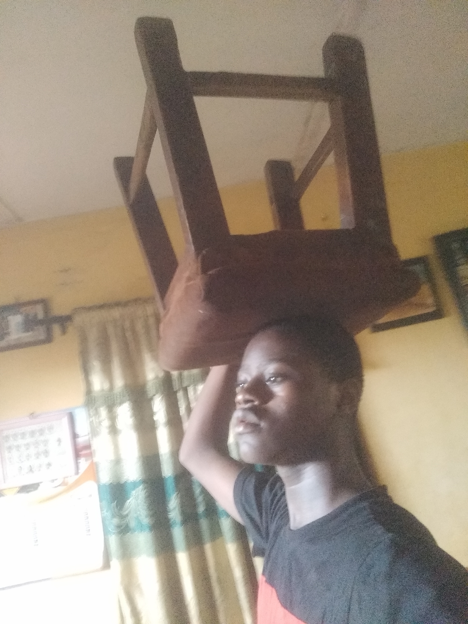 A young man stands indoors beneath a wooden chair balanced on his head. He wears a black t-shirt and gazes slightly upward and to his right. Behind him, a yellow wall is visible, adorned with framed pictures. Curtains hang near a window, and a white appliance, possibly a refrigerator, is partially visible. The location is Unknown, Unknown.