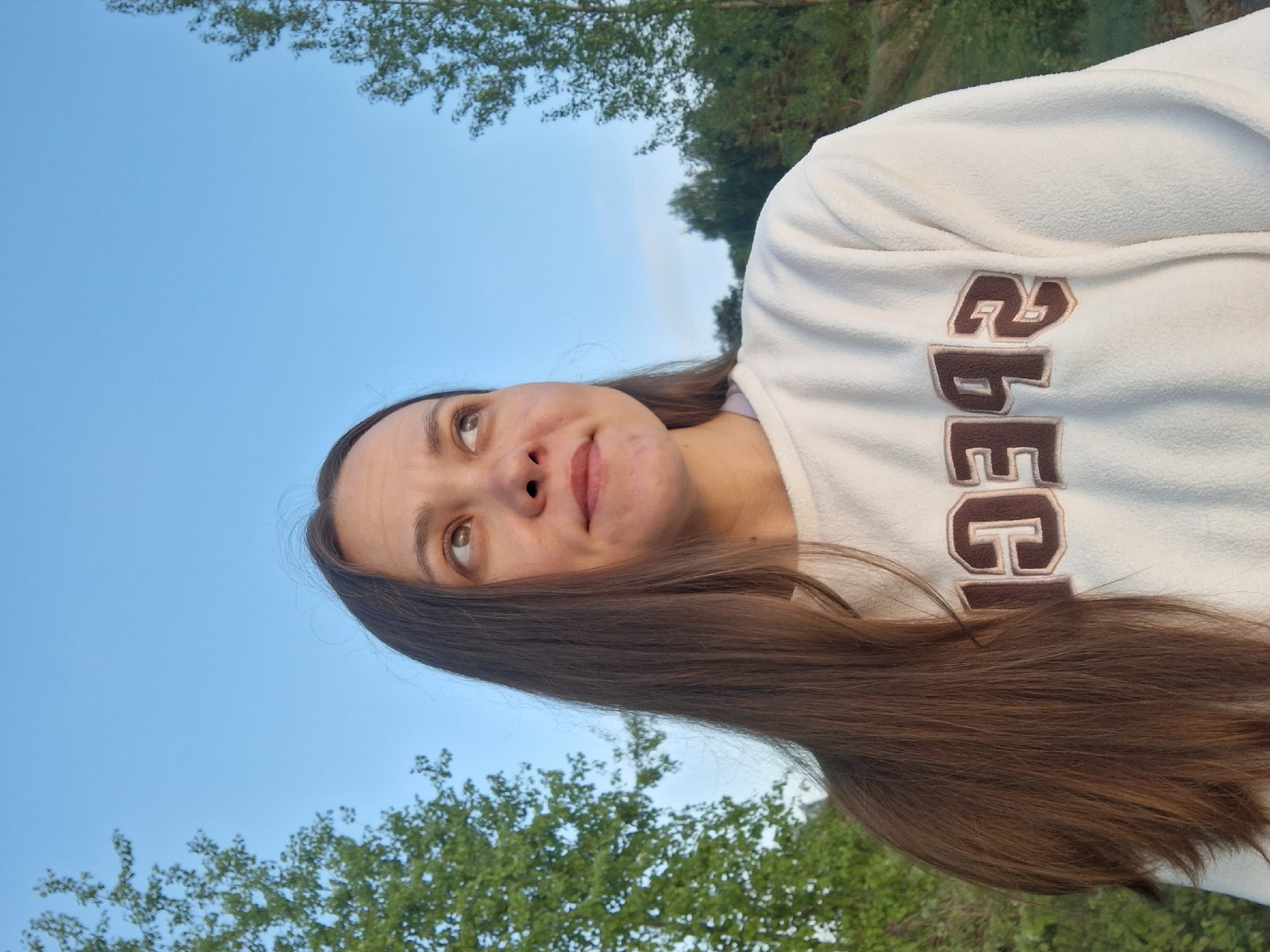 A woman with long brown hair and a cream-colored sweatshirt is looking upwards against a backdrop of a blue sky and trees. The sweatshirt has brown letters, but the word is not fully visible. The woman's face is tilted upwards, and she has a slight smile. The setting appears to be outdoors, with trees and vegetation visible in the background. The location, Chaikovsky, Russia, cannot be determined from the image alone.