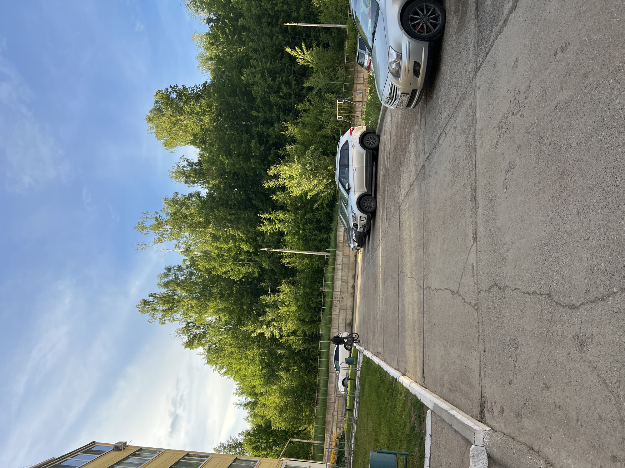 The scene depicts a street in Noviy, Russia. A silver sedan is parked next to a white SUV on an asphalt road with visible cracks. A person on a bicycle is near the white SUV. Lush green trees line the edge of the road, creating a backdrop to the vehicles. A fence separates the trees from a grassy area near a building. The sky is blue with scattered clouds.