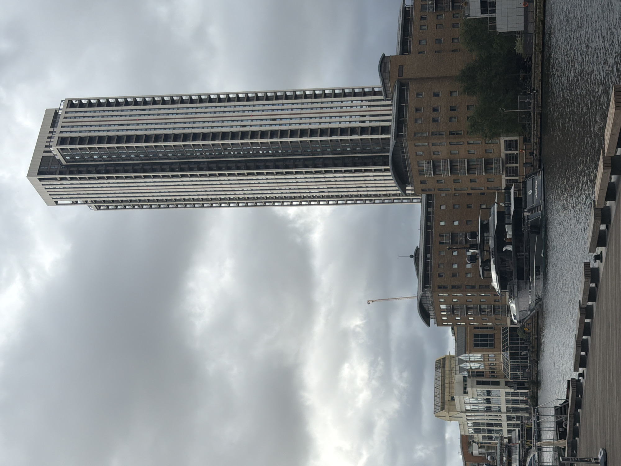 The image, correctly oriented, depicts a contemporary urban waterscape under an overcast sky. A towering, slender high-rise building dominates the left side of the frame, characterized by its numerous floors, horizontal bands of balconies or ledges, and alternating light grey and darker window sections.
To the right, a multi-story building with a more traditional, possibly brick-clad facade stands by the edge of a body of water, likely a canal or dock. This building features numerous windows and some curved architectural elements. In the mid-ground, a construction crane is visible against the cloudy backdrop, suggesting ongoing development.
The water, which appears calm with subtle ripples, fills the right-center of the image. Several boats are moored along the waterfront next to the brick building. One boat clearly displays the text "THE GREAT PERFUME" on its side. In the immediate foreground on the right, a wooden boardwalk or deck runs parallel to the water's edge, featuring dark-colored bollards or mooring posts. Below the brick building, another structure with a more modern, glass-fronted design is partially visible.
The scene is captured during the daytime under consistently grey, cloudy weather conditions, which casts a soft, even light over the landscape. The combination of modern high-rises, waterfront buildings, and boats strongly suggests a docklands or urban regeneration area in London, such as parts of Canary Wharf or the Royal Docks. There are no people visible, and the scene is static.