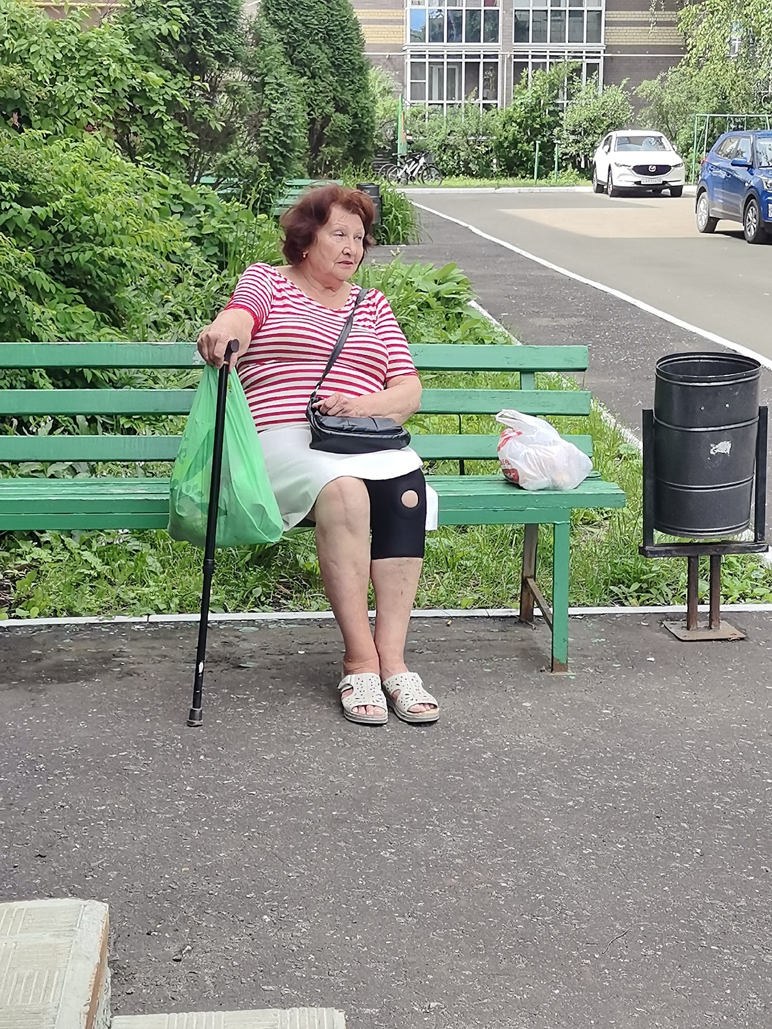 An elderly woman with short, reddish-brown hair sits on a green park bench in Nikolayevka, Russia. She is wearing a red and white striped t-shirt, a white skirt, and black compression sleeves on her legs, with white sandals on her feet. She holds a black walking cane in her right hand and has a green shopping bag on her left. A black purse is slung across her shoulder. Next to her on the bench is a clear plastic bag containing items. In front of the bench, to the right, is a black trash can. The bench is situated outdoors on a paved area with grass and trees in the background. A white car and a blue car are parked on a nearby road. A building with multiple windows is visible behind the trees.