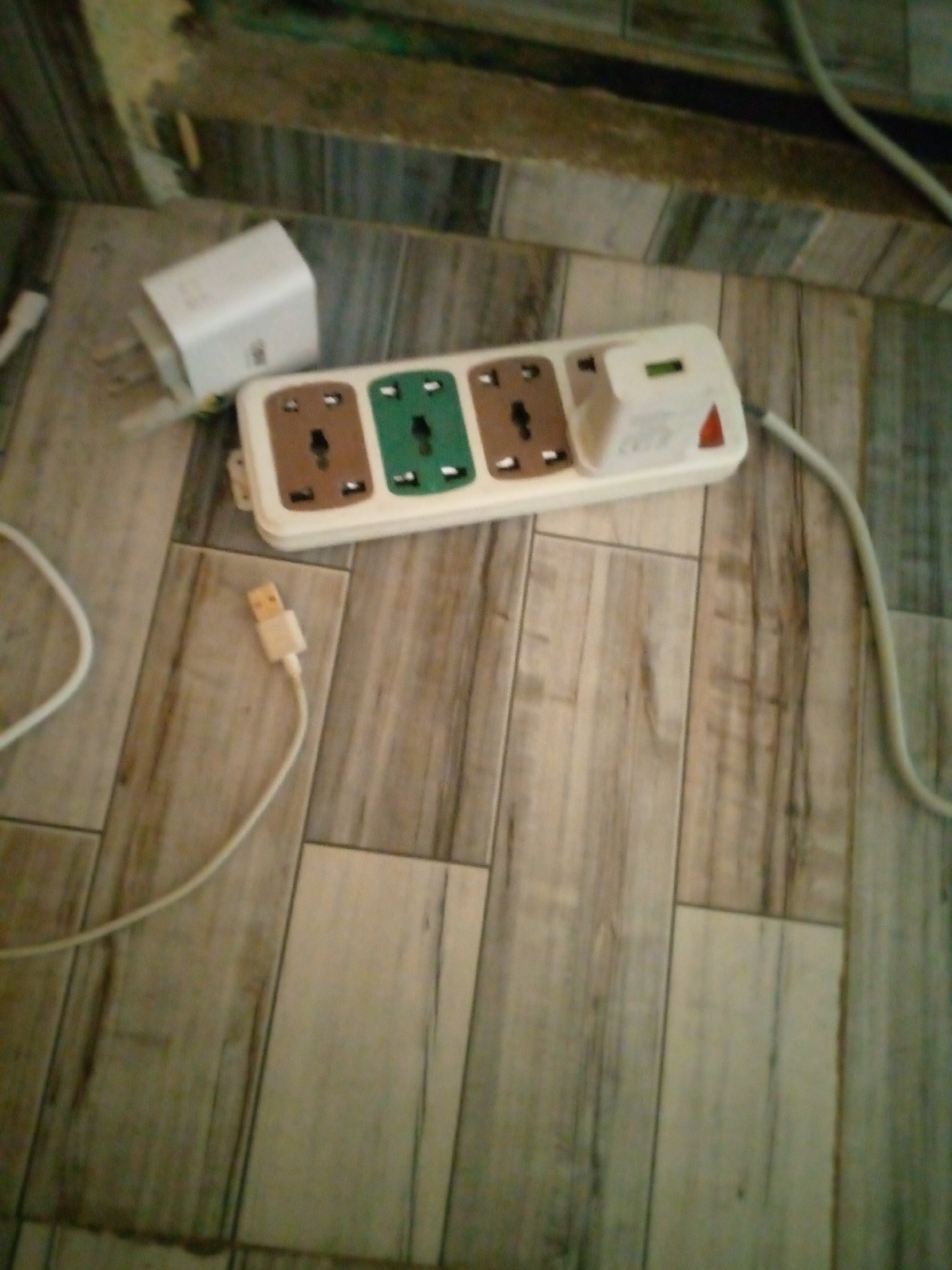 A power strip with three outlets is lying on a wooden floor. One outlet is brown, one is green, and one is brown. A white adapter is plugged into the green outlet. A white USB cable is plugged into the brown outlet. The floor is made of wooden planks. The wall behind the power strip is green and brown. The image was taken in Jalingo, Nigeria.