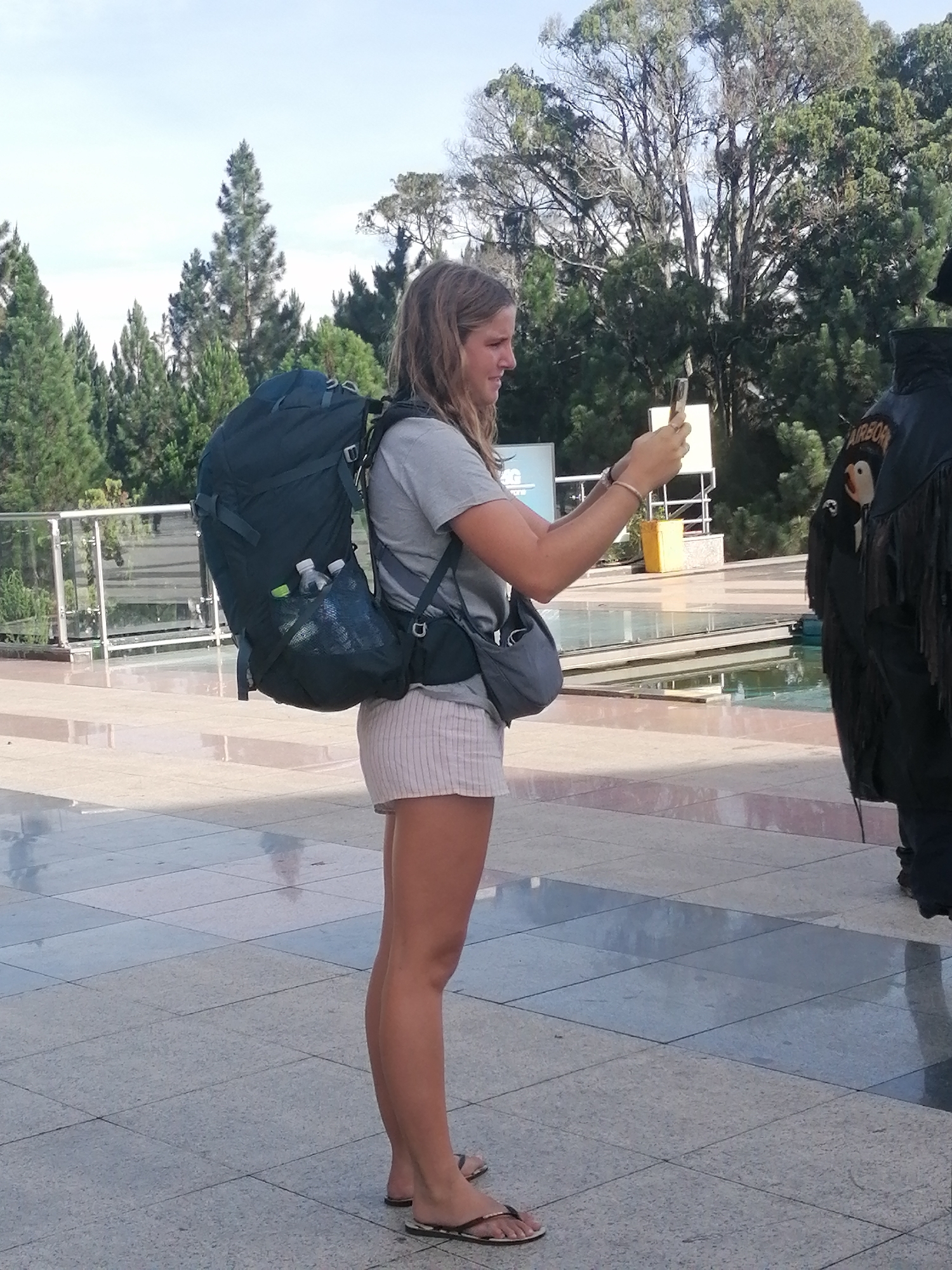 The image shows a young woman with long blonde hair, wearing a grey t-shirt, striped shorts, and flip-flops, standing outdoors. She is carrying a large dark blue backpack. In her hands, she holds a mobile phone, appearing to take a picture or video. To her right, another person is visible from the waist down, wearing a dark garment with fringed sleeves and an "AIRBORNE" patch. The background features trees, a body of water, and a paved walkway. The scene is likely set in Da Lat, Vietnam, suggested by the distinctive architecture and vegetation.