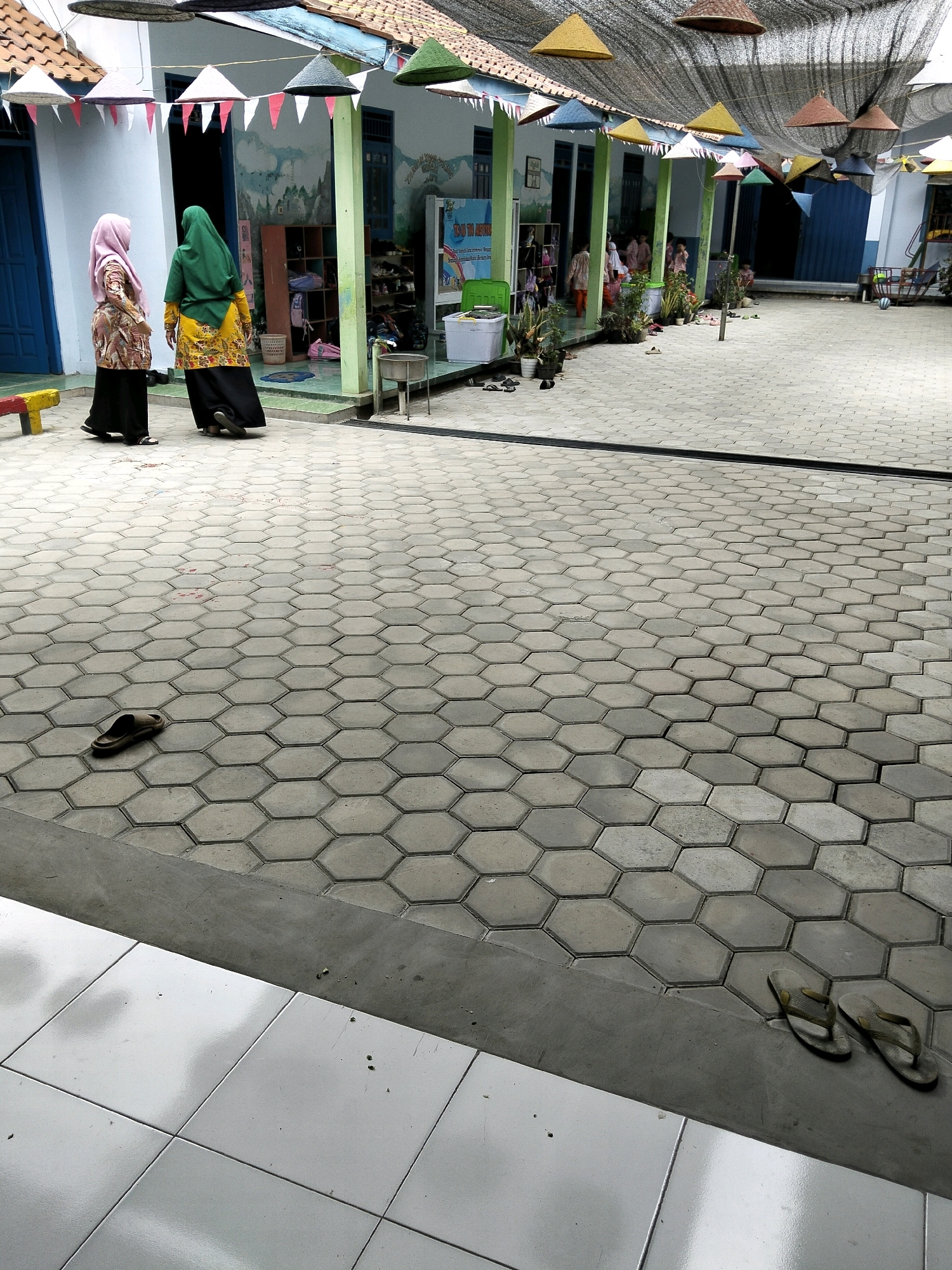 The image captures a courtyard area of a building, likely a school or community center, in Krajan Lor, Indonesia. The ground is paved with hexagonal concrete tiles. In the foreground, a pair of dark-colored flip-flops rests on the edge of the tiled area, next to a section of smooth white tiles. Further into the courtyard, another single flip-flop is visible on the hexagonal tiles.
The building's exterior features white walls with blue doors and trim. Colorful triangular flags and conical, woven shades are strung across the covered walkway. Several potted plants are positioned along the building's facade.
Two individuals, both wearing headscarves and modest clothing, are walking away from the camera in the middle ground. One wears a pink headscarf and a patterned tunic over black pants. The other wears a green headscarf and a yellow, patterned tunic over black pants. They appear to be heading towards the building.
In the background, more people, including children, can be seen entering or inside the building. There are shelves with items, bins, and a poster visible through an open doorway. The overall scene suggests a daytime setting in a semi-outdoor educational or communal space.